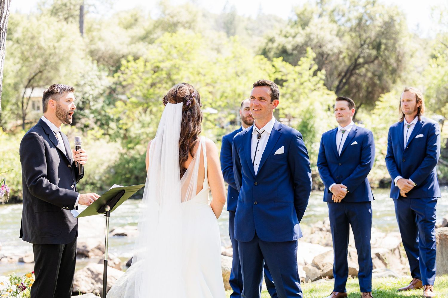Wedding party in navy blue suits and white dresses gather outdoors for candid photos in a natural setting.