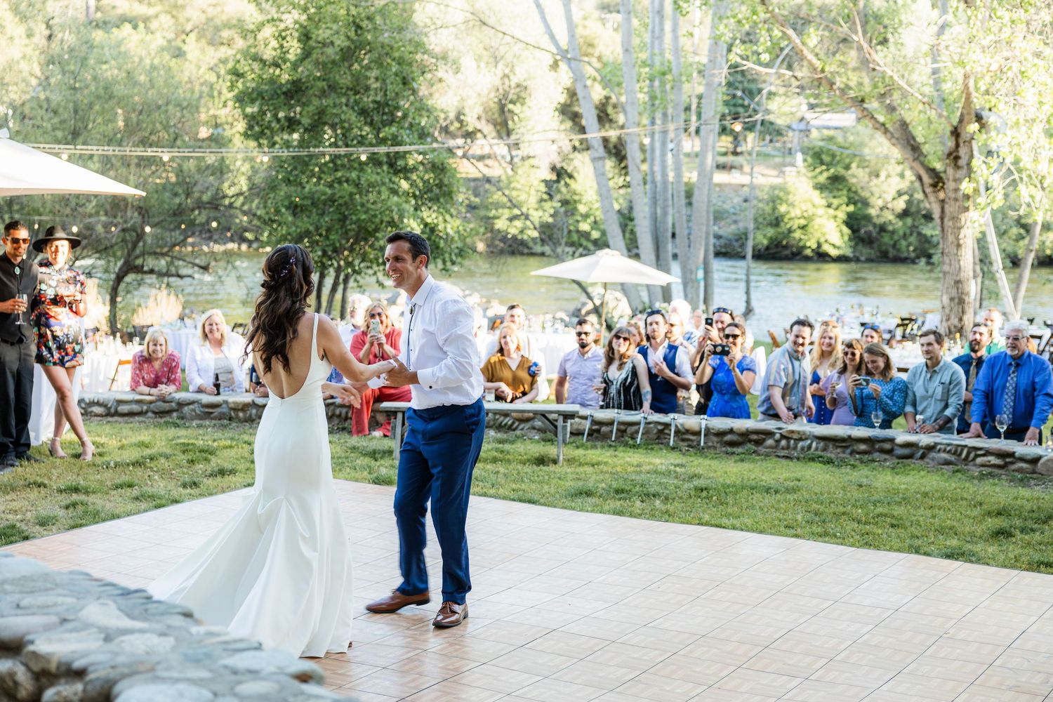 Wedding ceremony guests seated outdoors on white chairs watch as couple walks down grassy aisle at sunset.