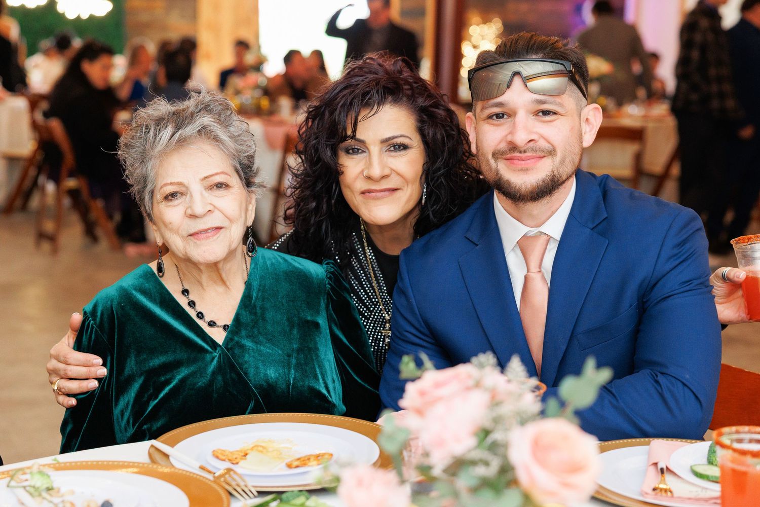 Three people smile together at a formal event with table settings and pink flowers in the foreground.