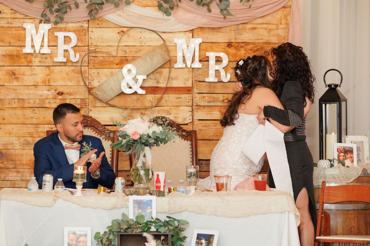 Wedding reception guests seated at rustic wooden tables with white decor and greenery in an intimate indoor venue.