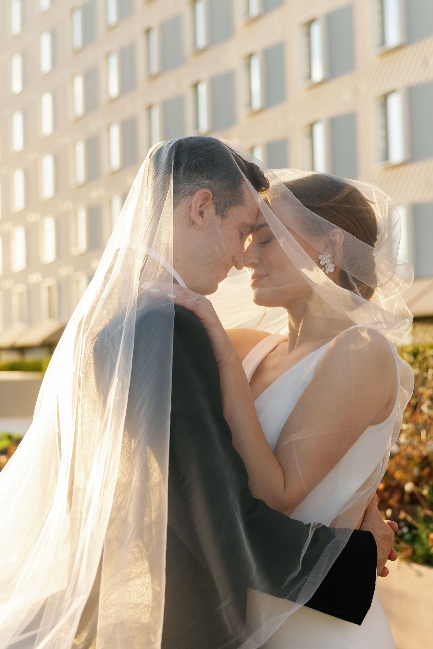 A romantic wedding couple embraces during sunset with a flowing veil and elegant attire.