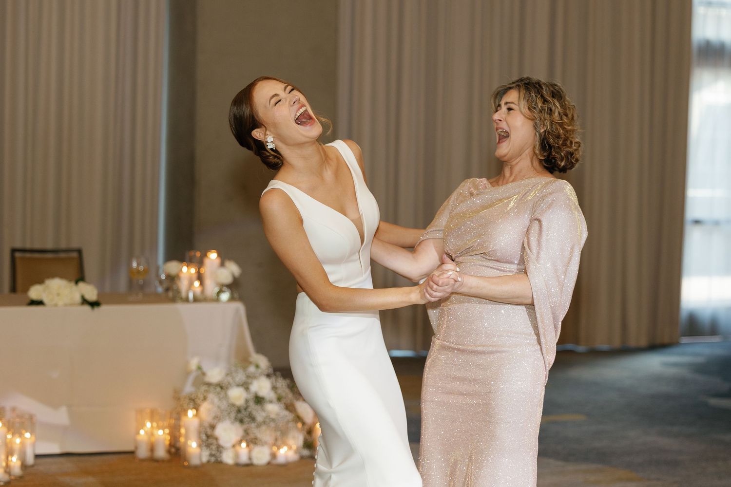 Two people in formal attire dance and laugh together in a bright indoor venue with candles in the background.