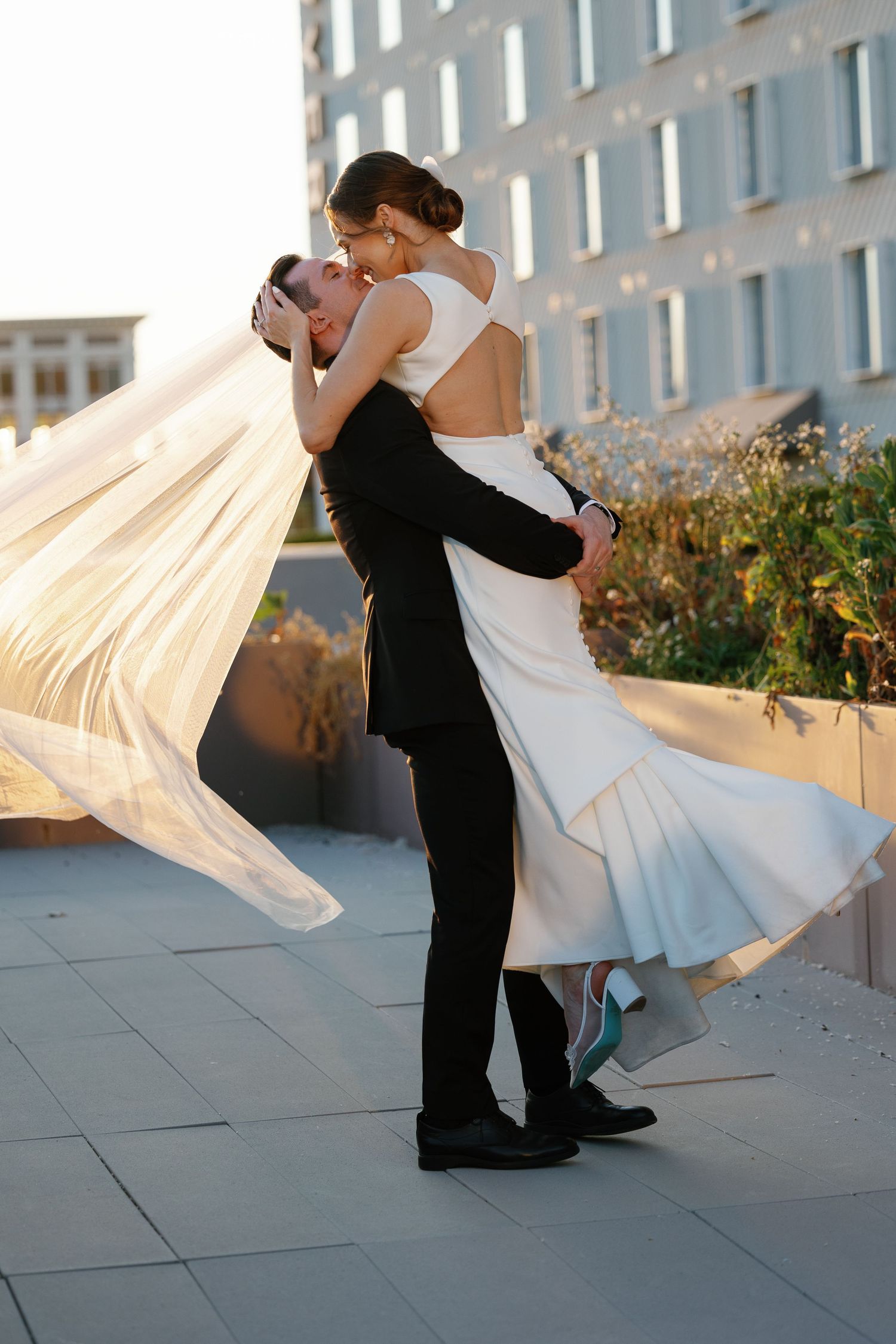 A couple dances together at sunset, with the bride's white dress flowing gracefully in the breeze against a modern building backdrop.