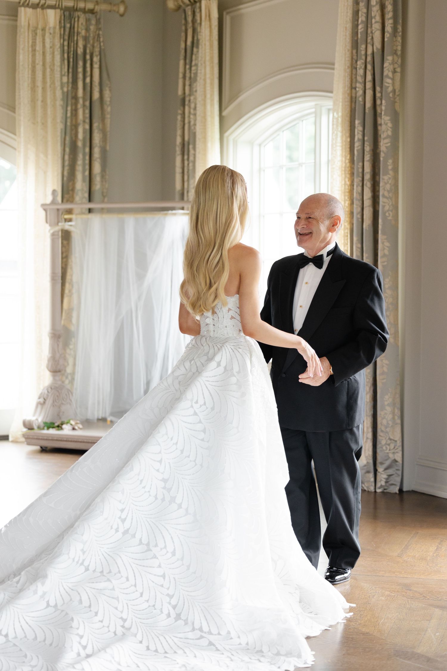 A couple in formal wedding attire dances together in an elegant room with large windows and flowing curtains.