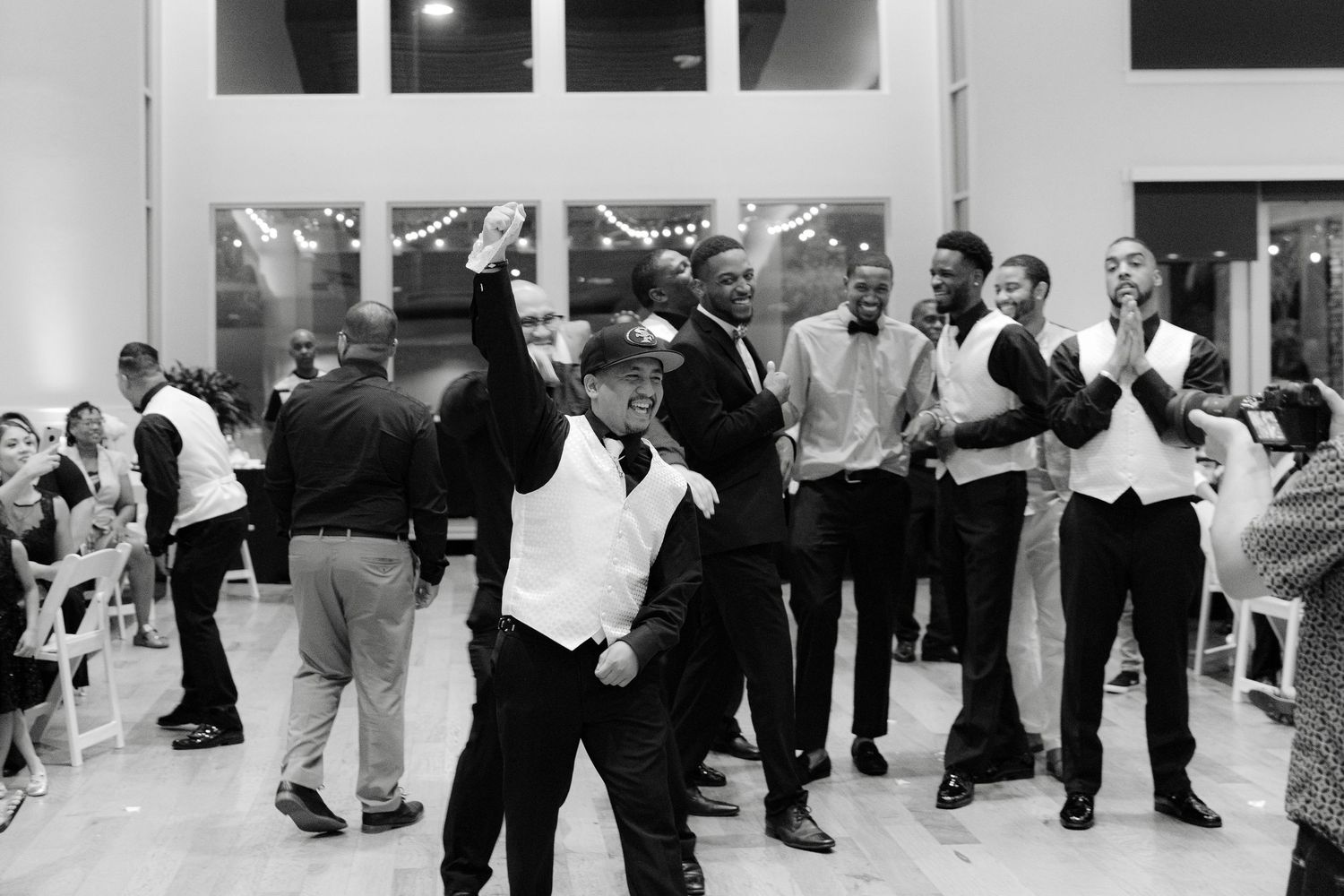 Black and white photo of people dancing and celebrating at a lively party or reception in an indoor venue.