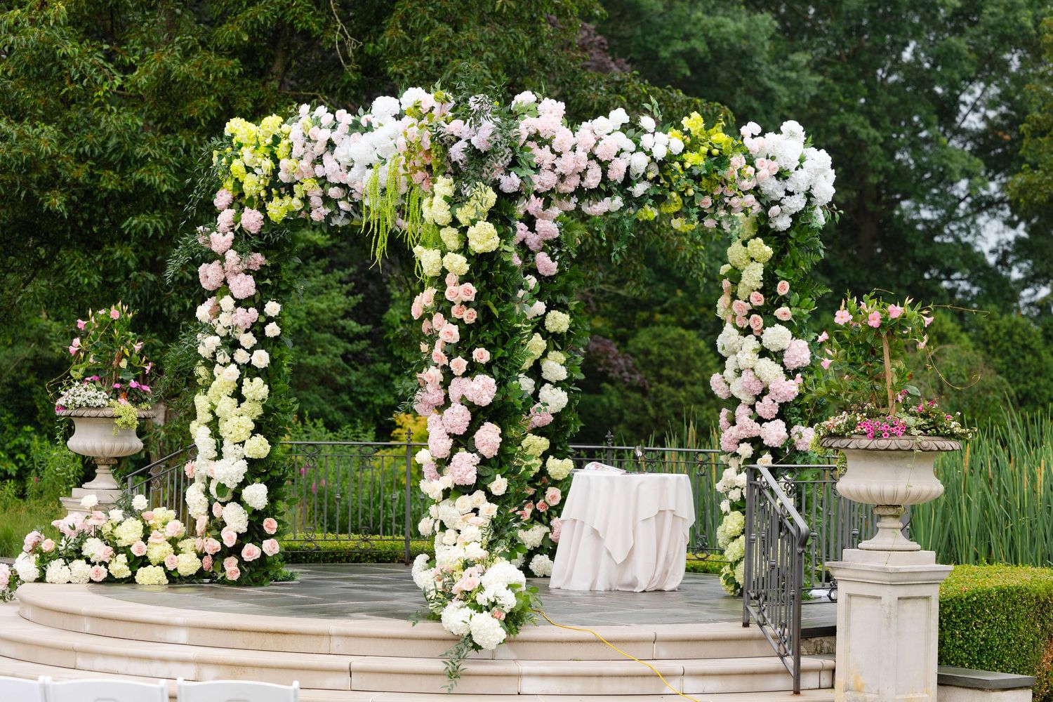 Elegant white floral archway decorations adorn a garden wedding ceremony space with stone pillars and greenery.