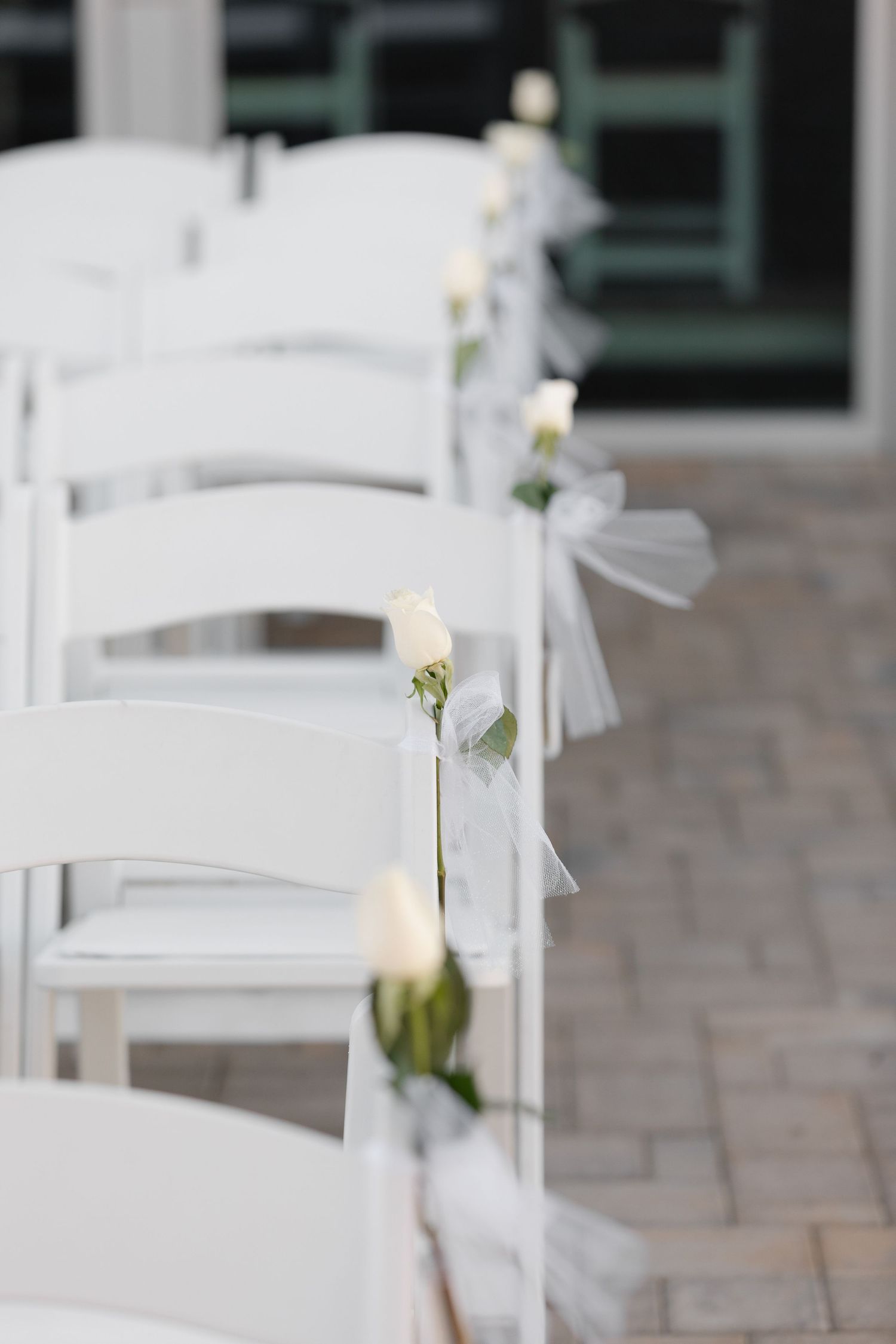 White folding chairs with delicate floral decorations line up for an outdoor wedding ceremony on stone pavement.
