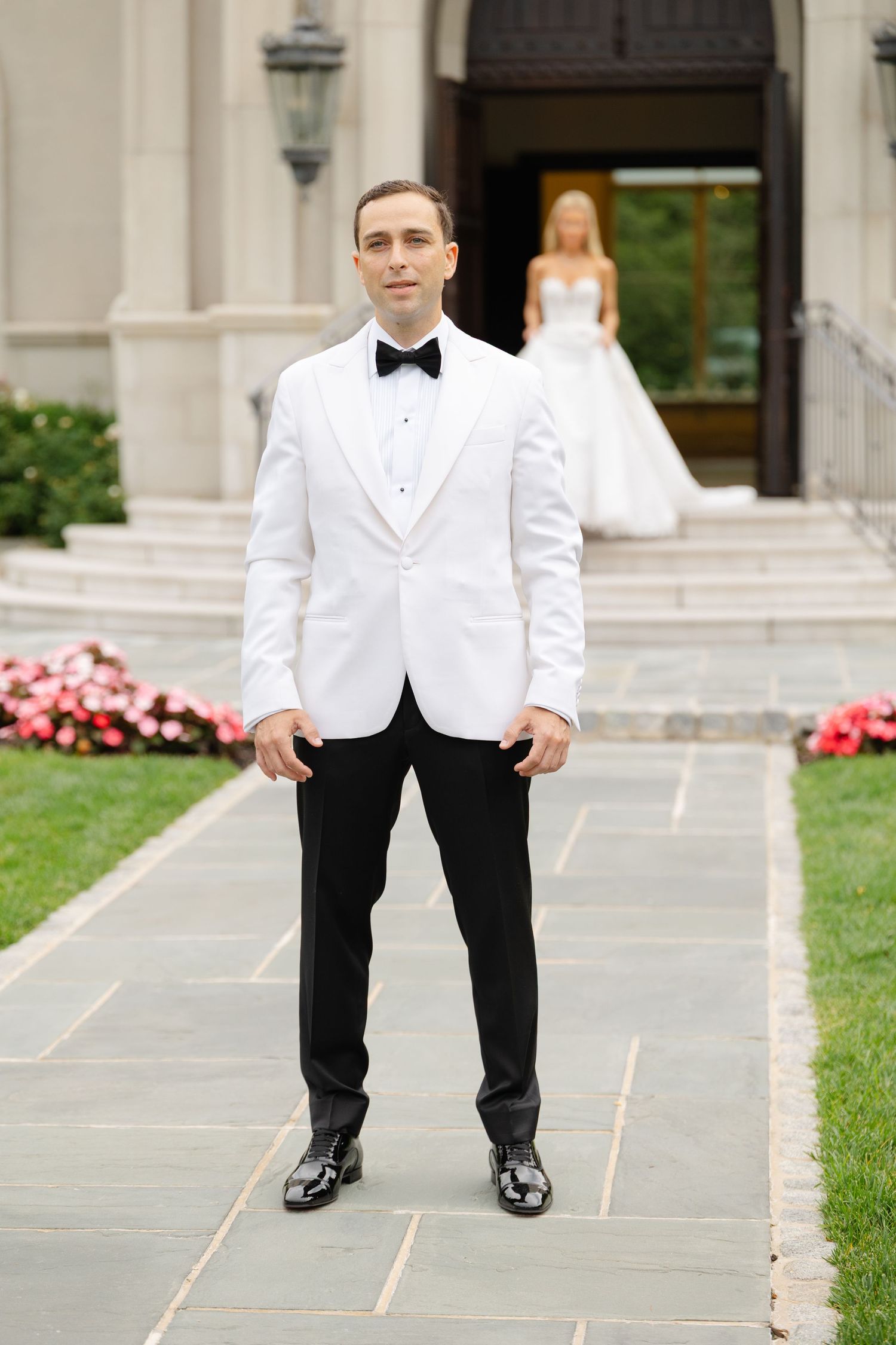 A person in a white dinner jacket and black bow tie stands on stone steps near pink flowers and greenery.