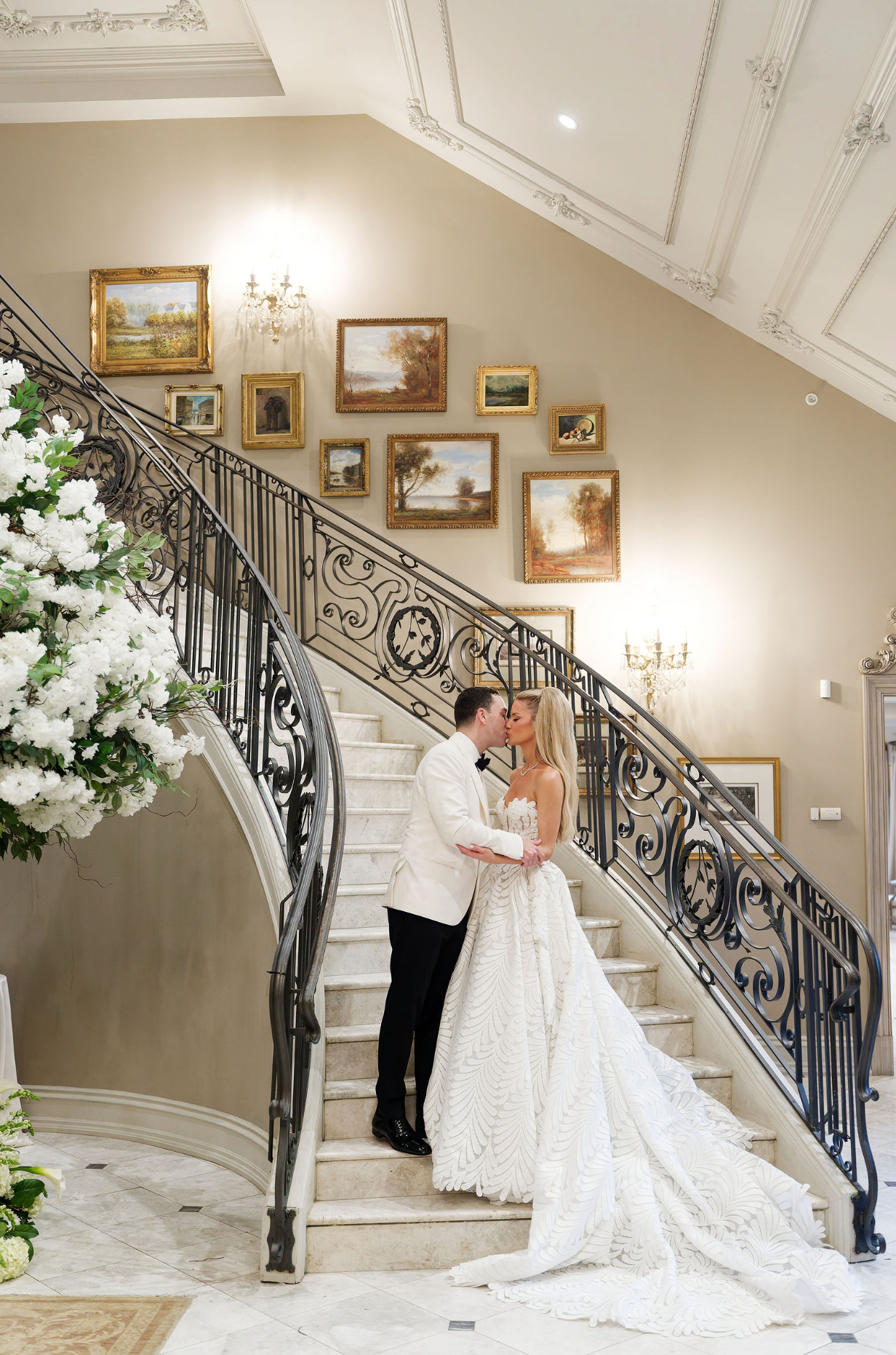 Elegant wedding scene on a grand staircase with ornate iron railings and framed artwork adorning the walls.