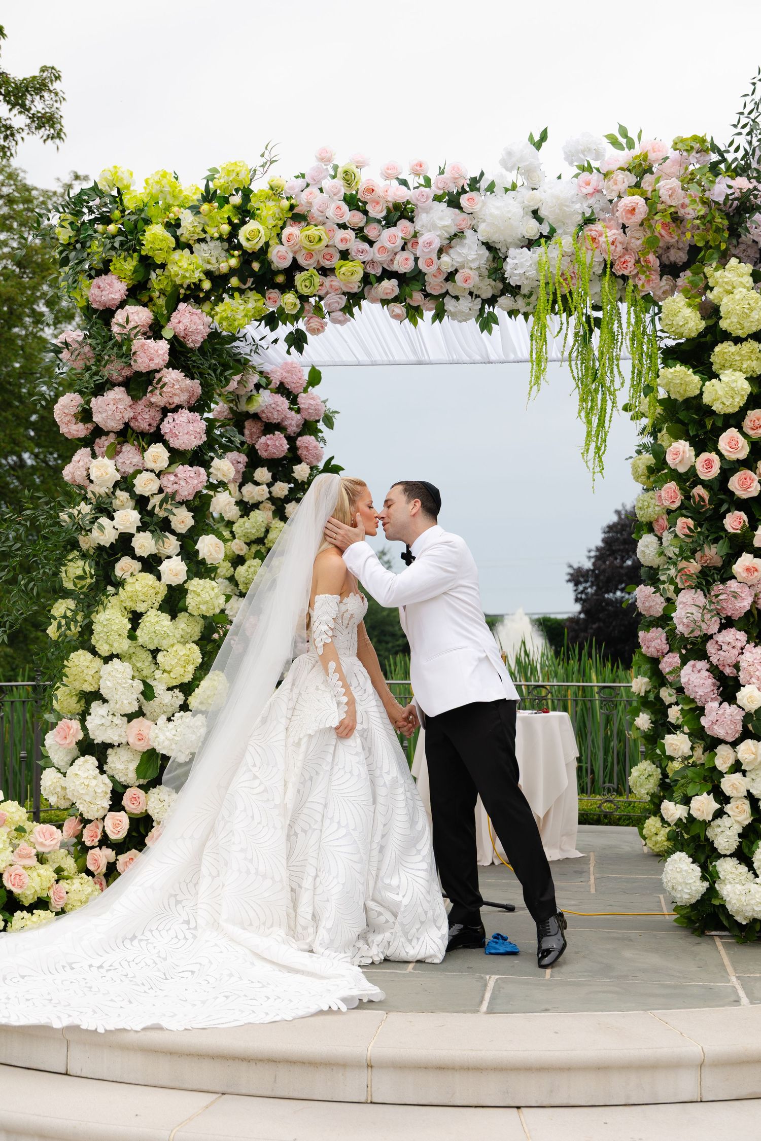 A romantic wedding ceremony moment under an elegant floral arch with pink and white roses and cascading greenery.