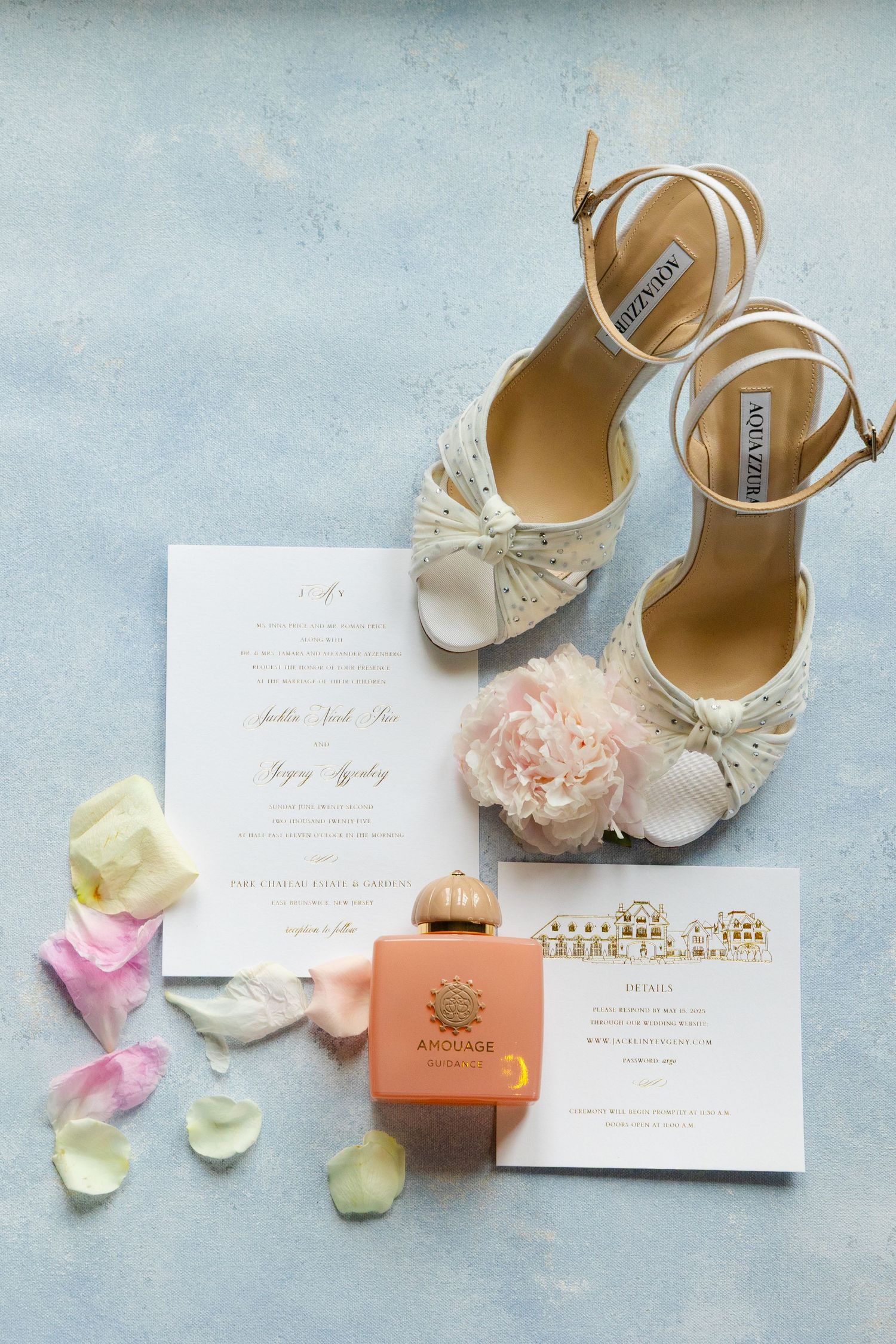 Elegant bridal shoes with floral details arranged with wedding invitations, rose petals, and peach-colored gift box on blue surface.