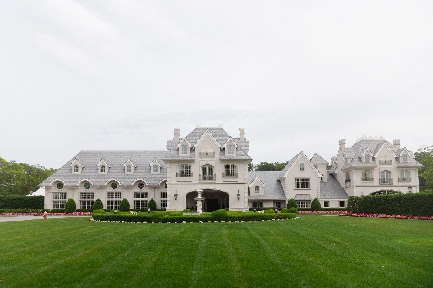 Luxurious white mansion with French chateau architecture sits on a manicured green lawn under cloudy skies.