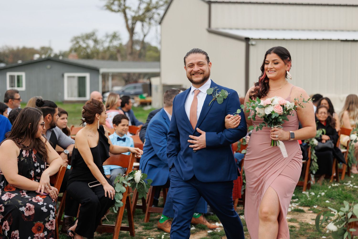 A joyful couple walks down the aisle at an outdoor wedding ceremony while guests look on from their seats.