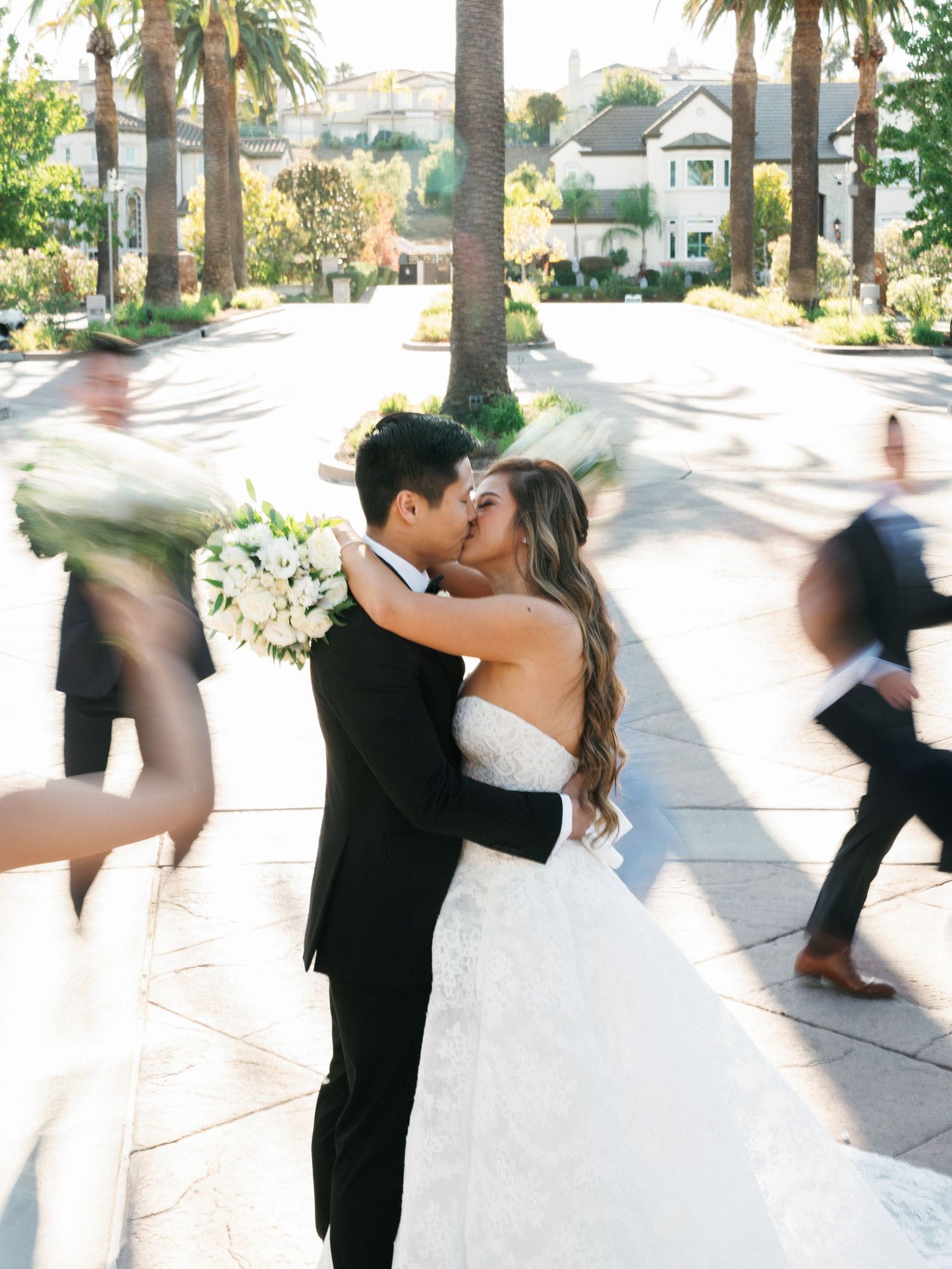 A romantic moment captured between newlyweds with palm trees and motion-blurred guests in the background.