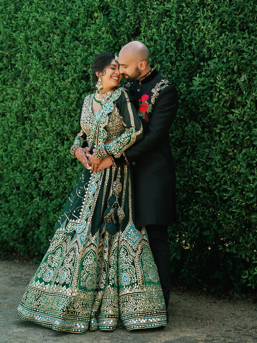 Traditional Indian bridal couple poses against lush green hedge backdrop in ornate wedding attire.