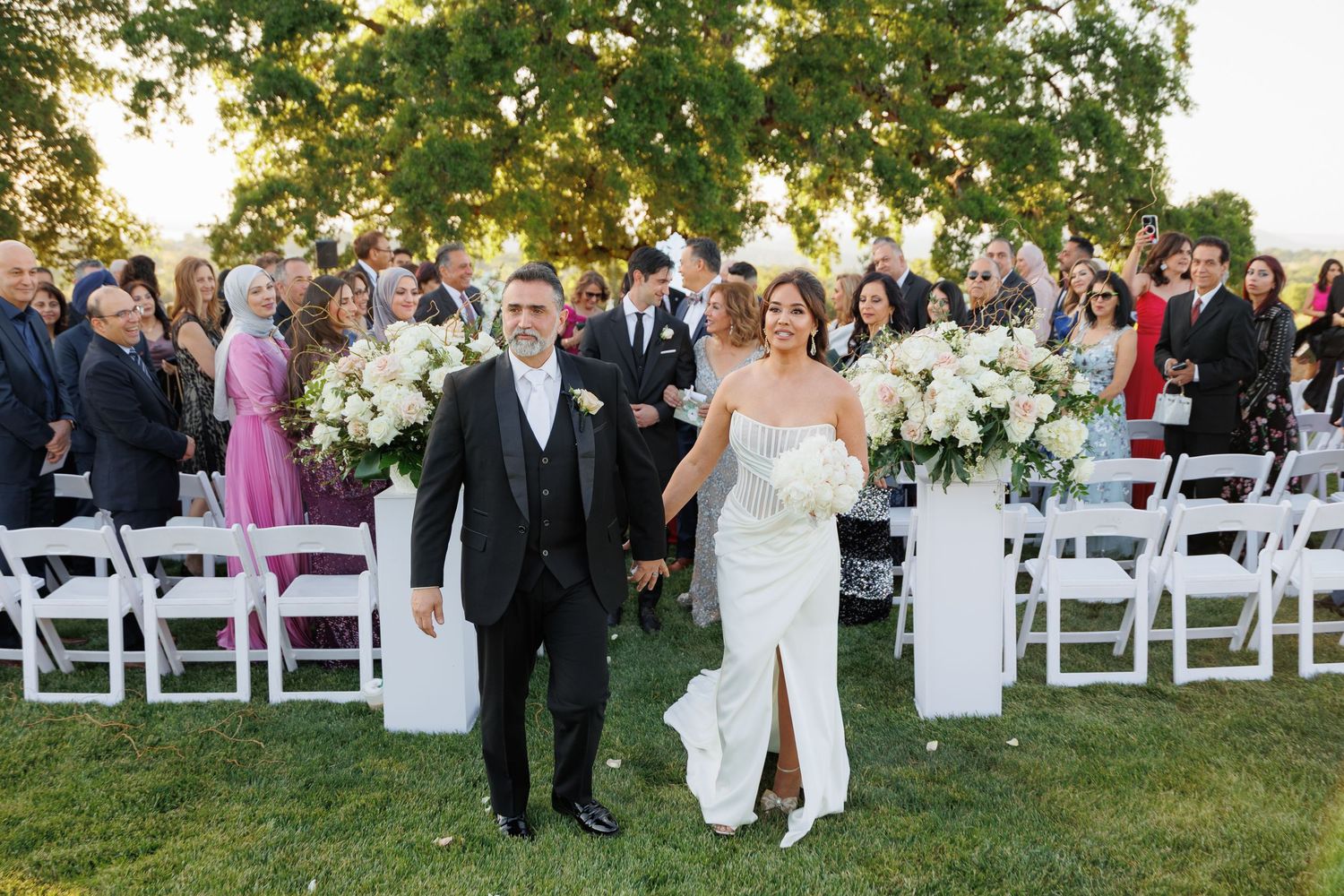 Outdoor wedding ceremony setup with white chairs arranged on a grassy lawn under trees during golden hour.