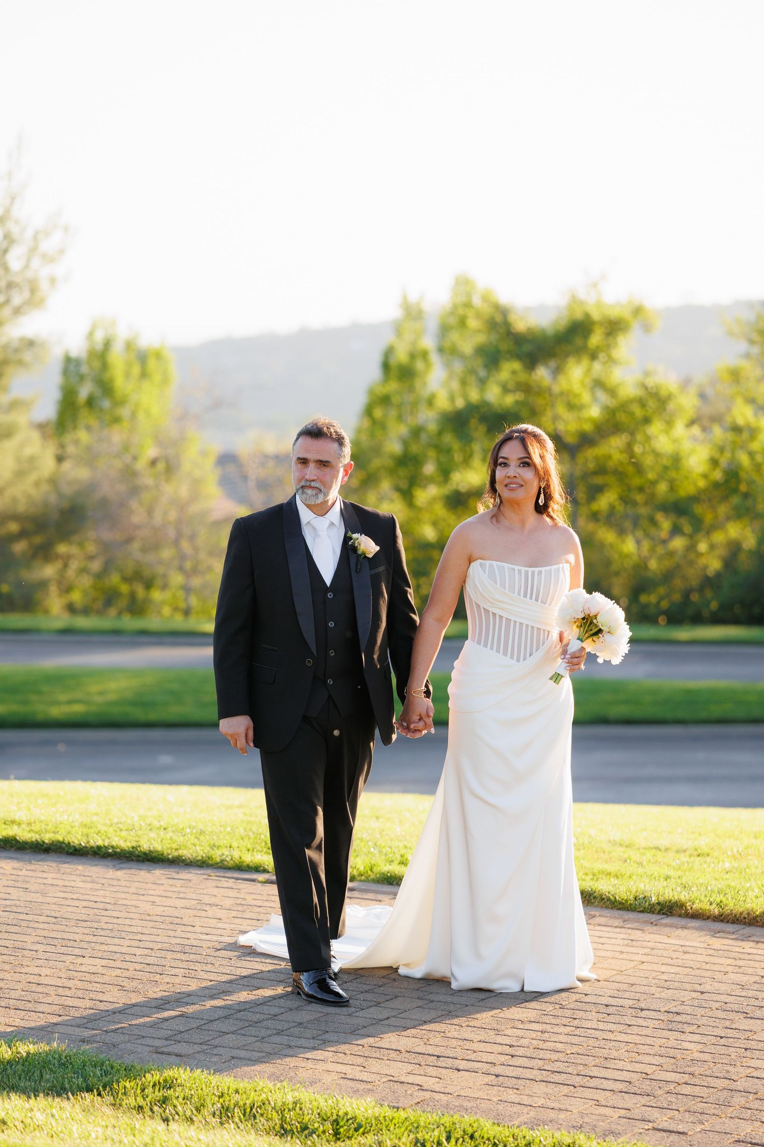 A couple walks together on a paved path during their outdoor wedding ceremony with mountains and trees in the background.