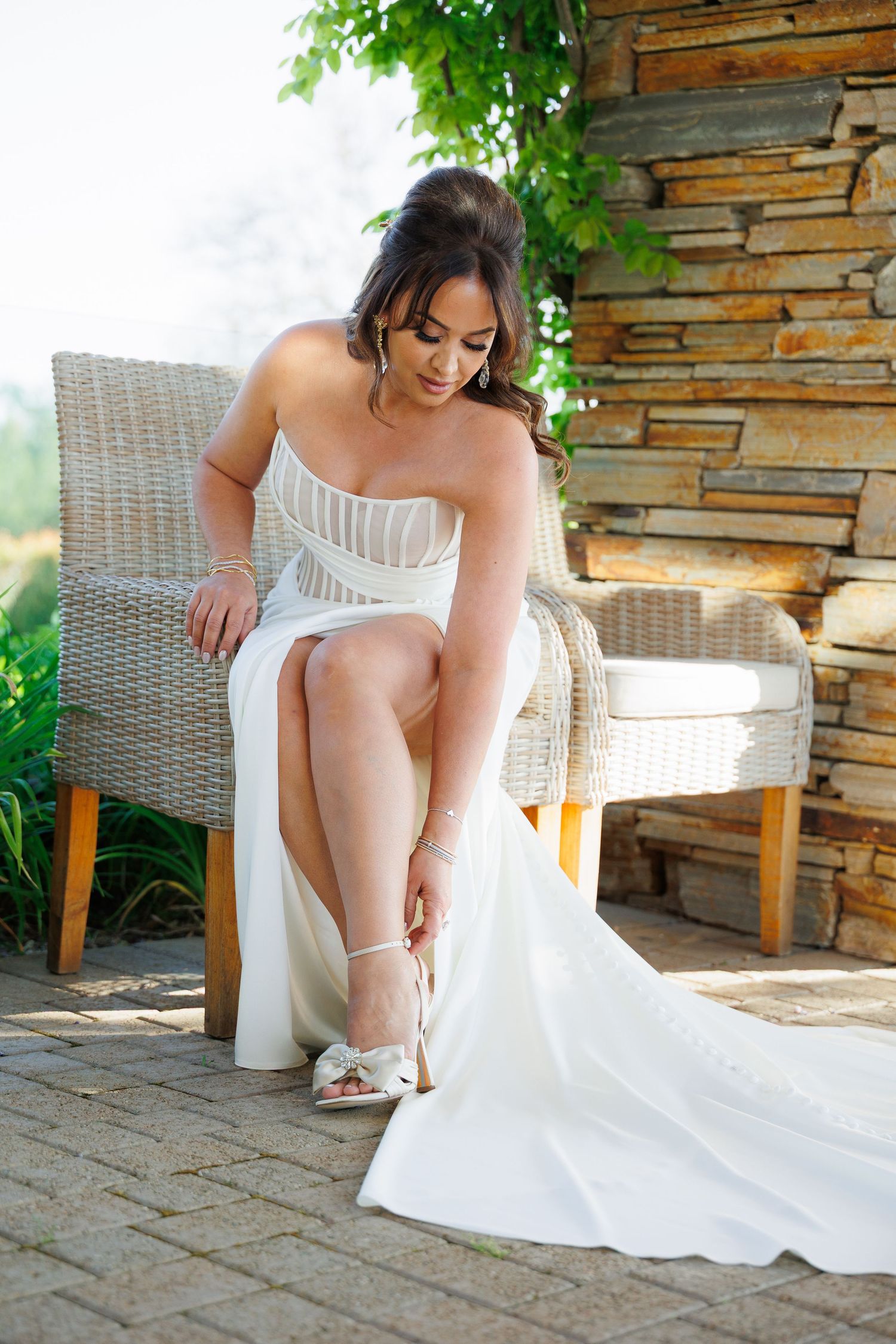 Bride in white dress adjusts her shoes while sitting in wicker chair on stone patio.