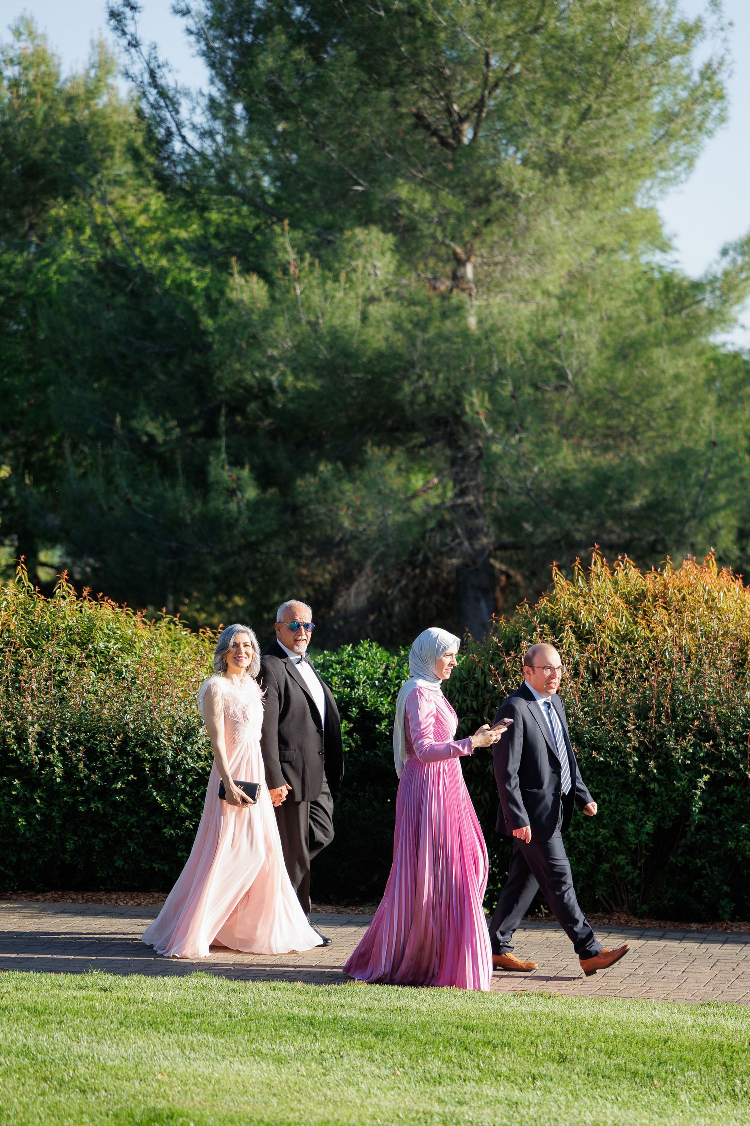 Wedding guests in formal attire including pink and blush gowns walk along a garden path on a sunny day.