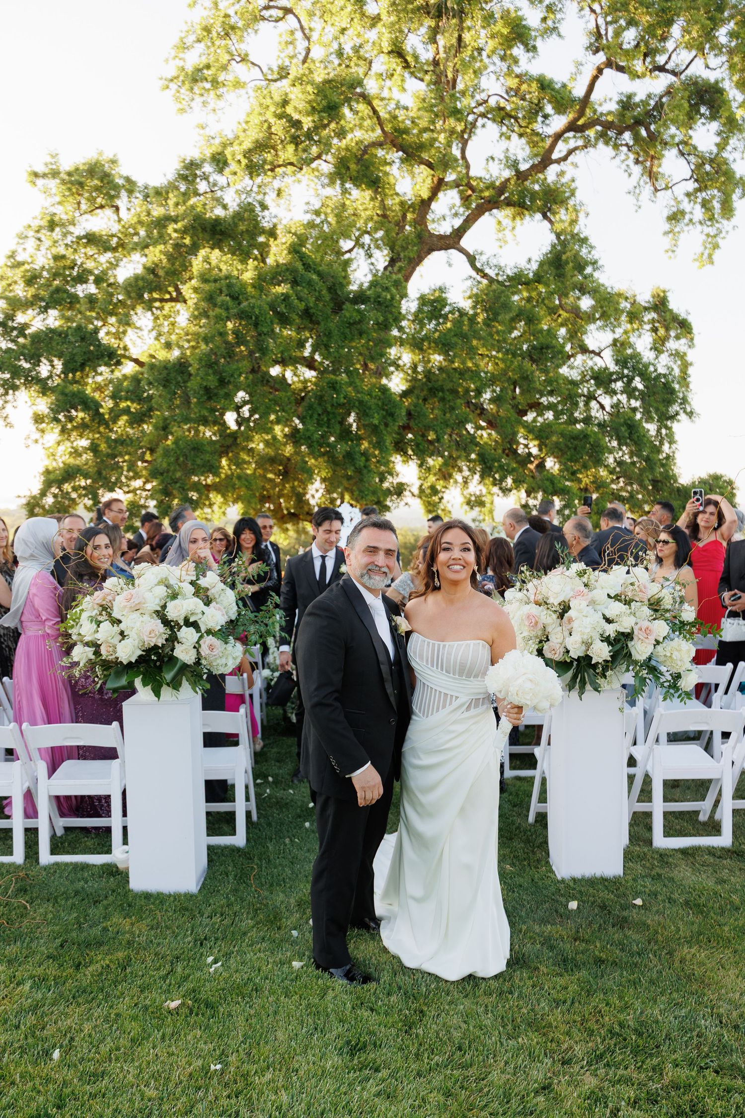 Wedding guests gather on a grassy lawn with white chairs and floral arrangements during an outdoor ceremony.