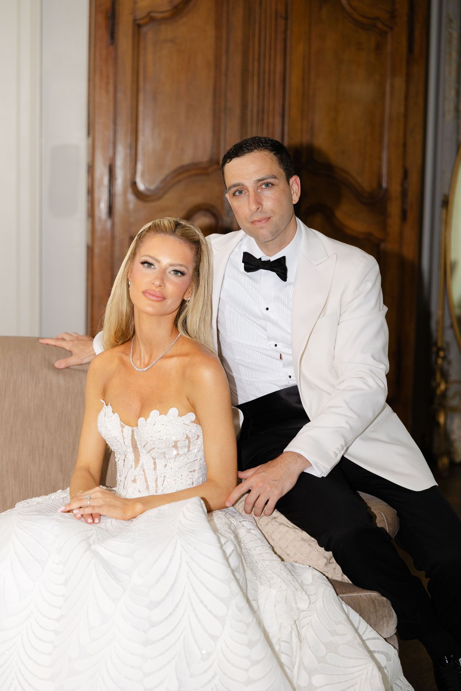 A bride in a white corset gown poses with a groom in a white tuxedo jacket against an elegant wood-paneled wall.