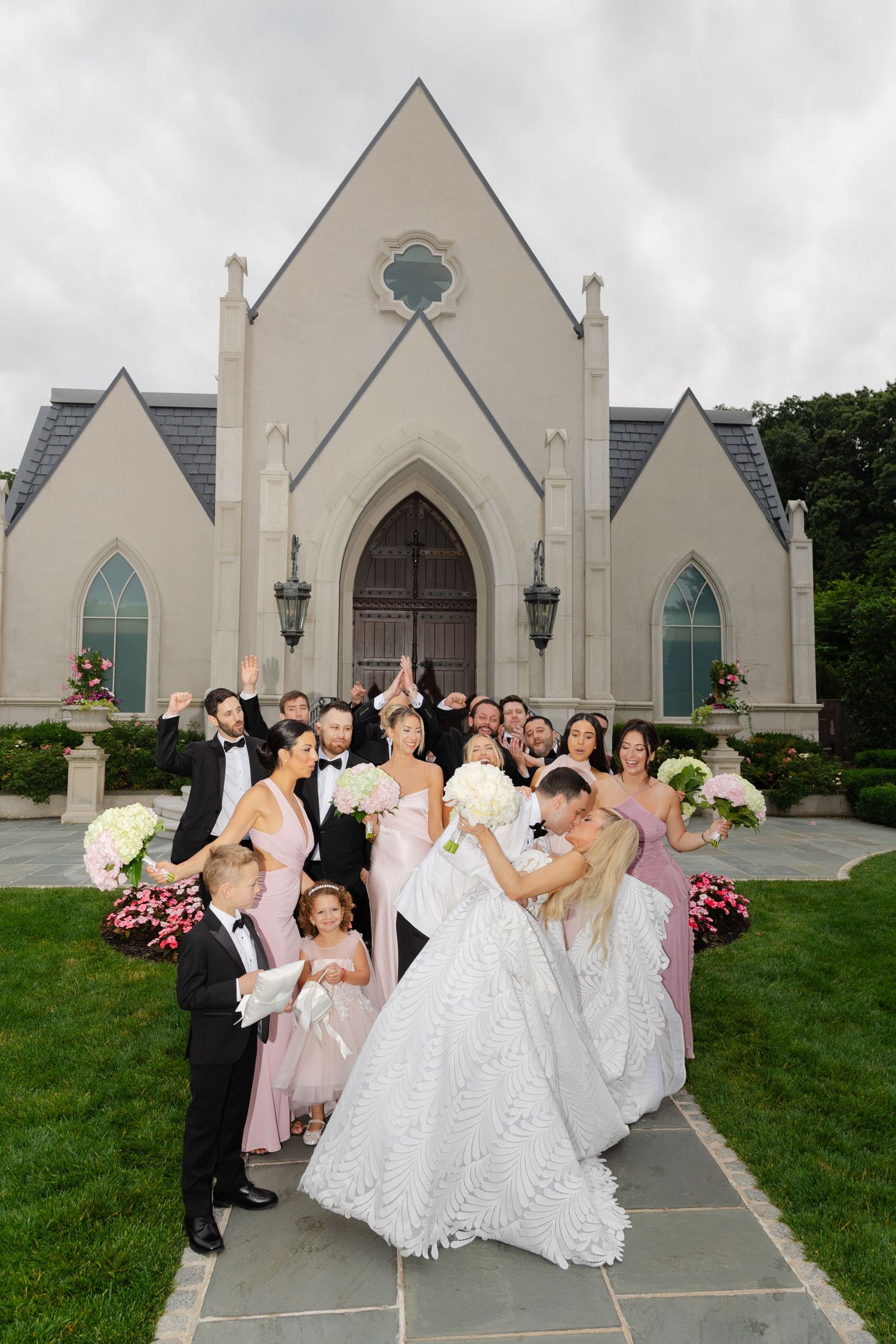 Wedding party poses for group photos outside a beautiful gothic-style church with pointed arches and white facade.