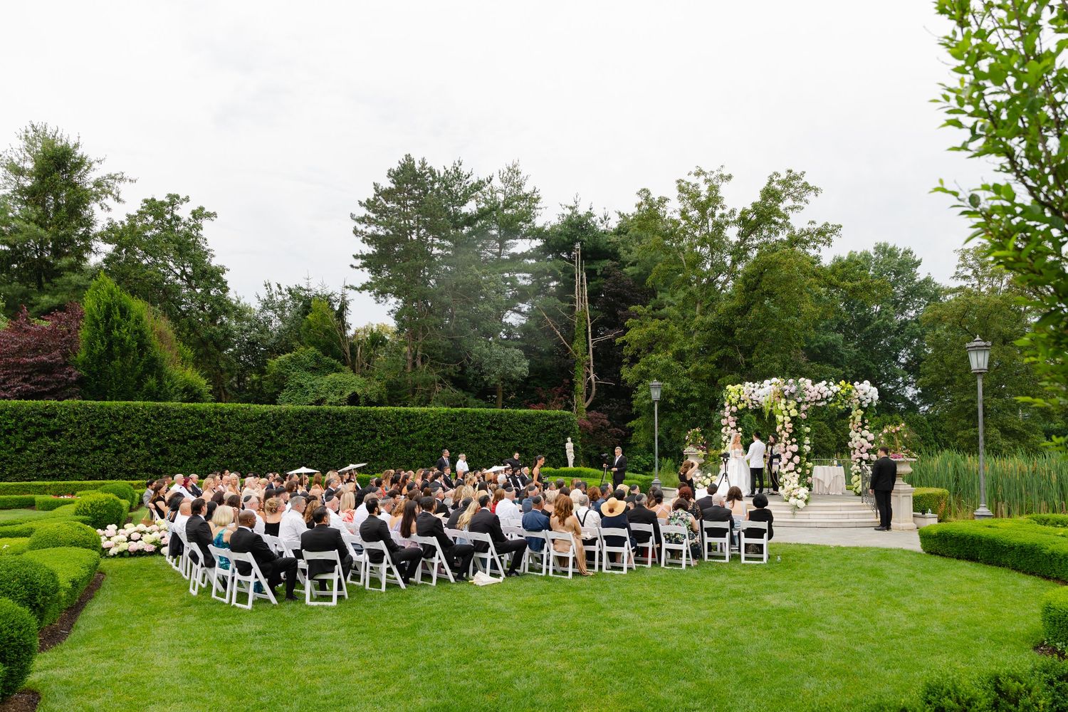 Wedding ceremony guests seated in white chairs on a lush green lawn surrounded by trees and hedges at an outdoor venue.