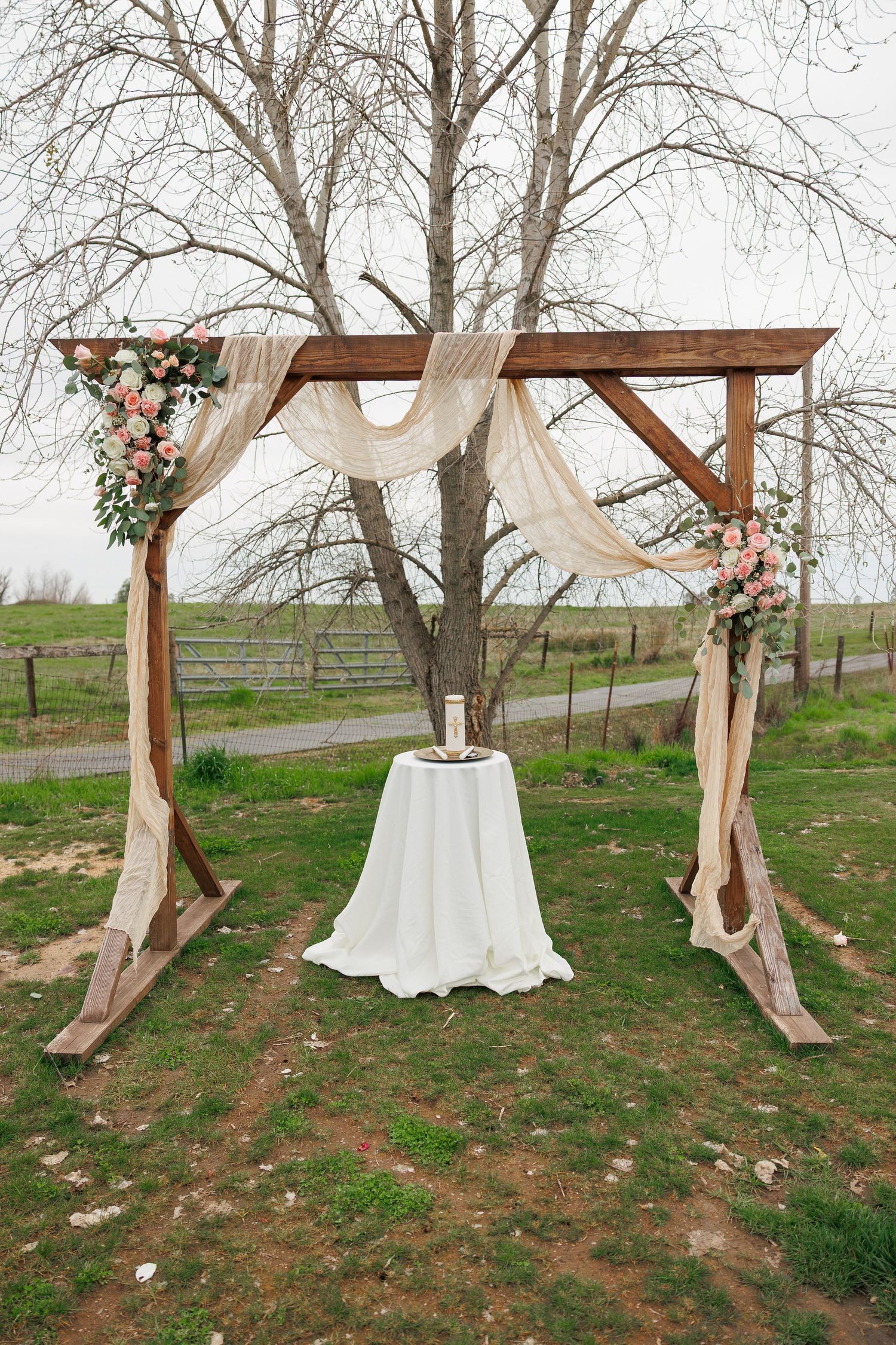 Rustic wooden wedding arch decorated with white fabric draping and pink floral arrangements in a rural outdoor setting.