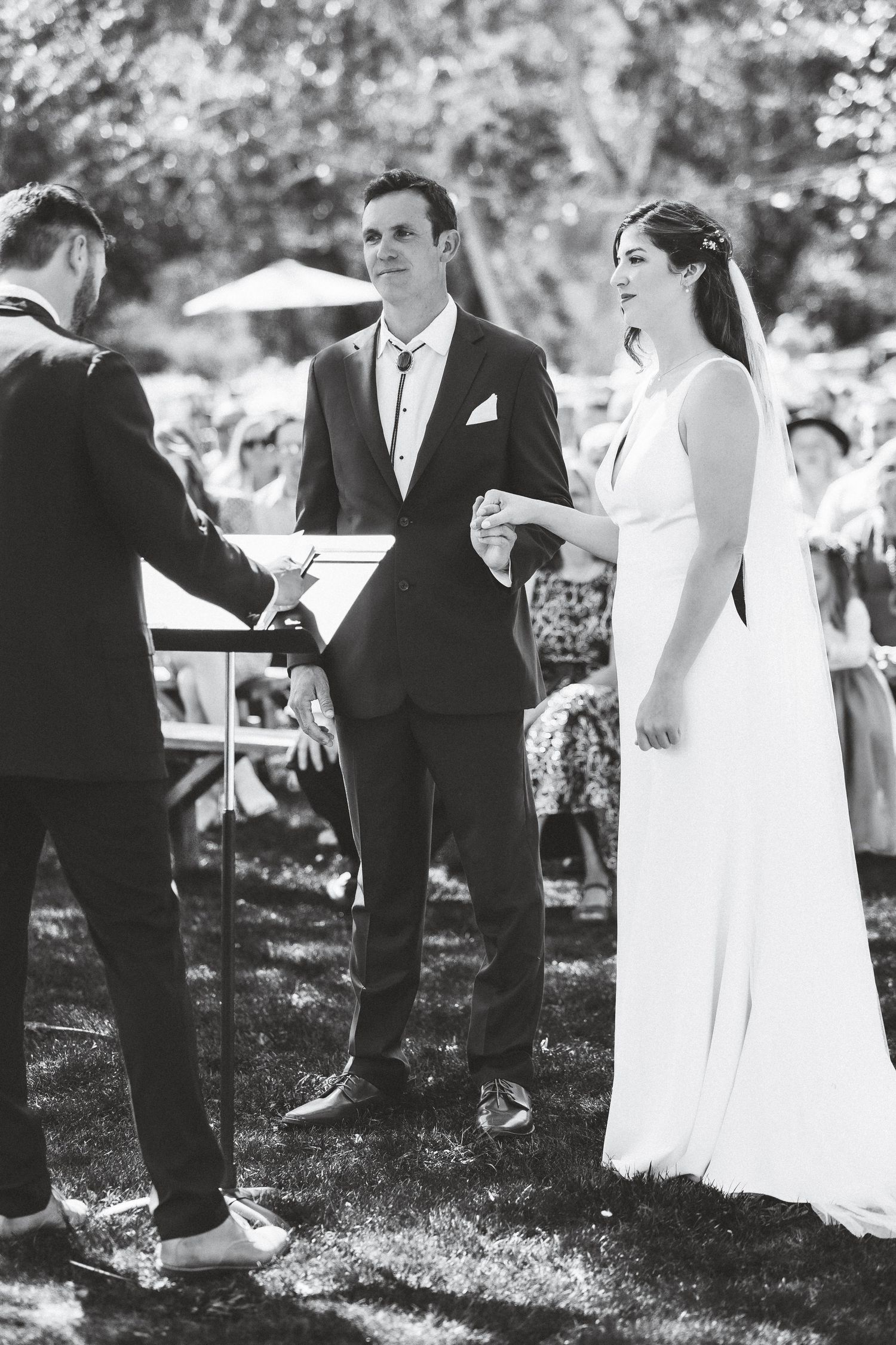 A black and white photo of a wedding ceremony taking place outdoors with an officiant and couple exchanging vows.