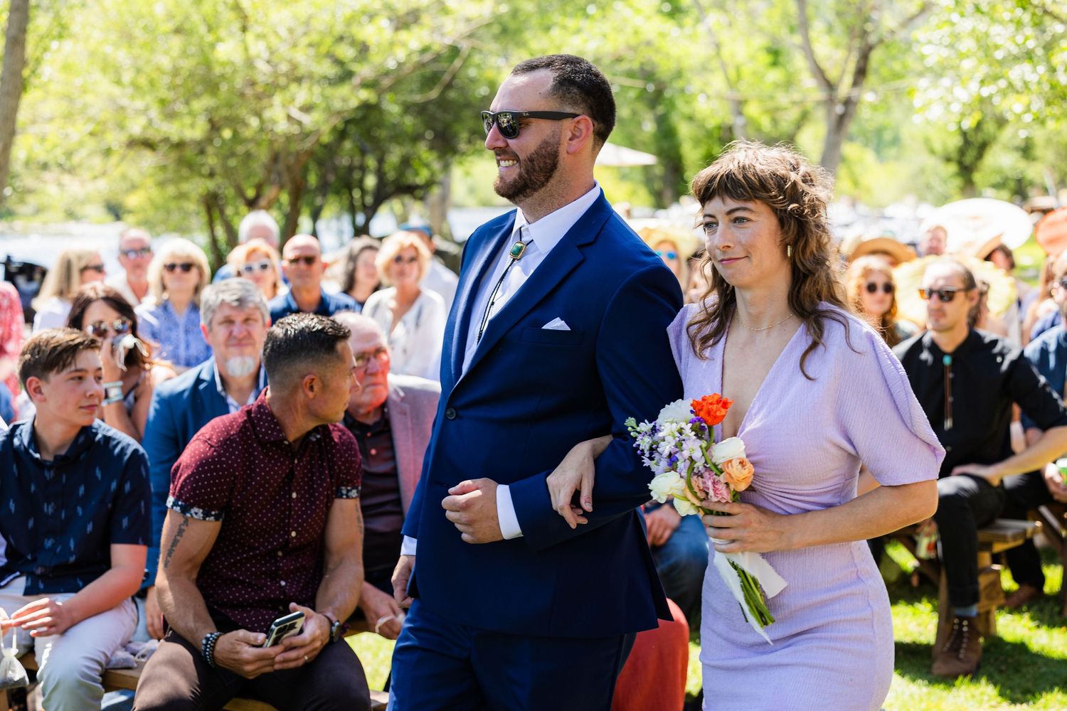 Wedding guests sitting outdoors in a garden setting laugh and smile during a sunny celebration.