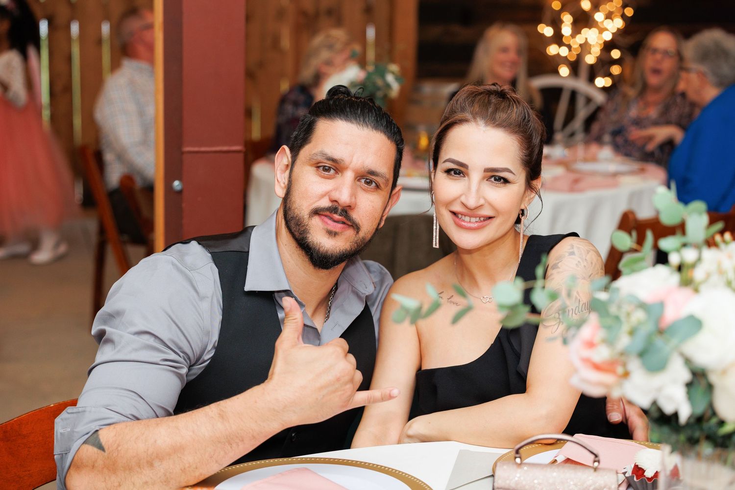 A couple smiles together at an elegant dinner reception with floral centerpieces and twinkling lights in the background.