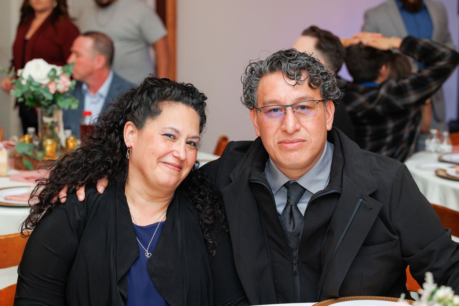 Two people in formal attire smile and chat while seated at a social gathering.