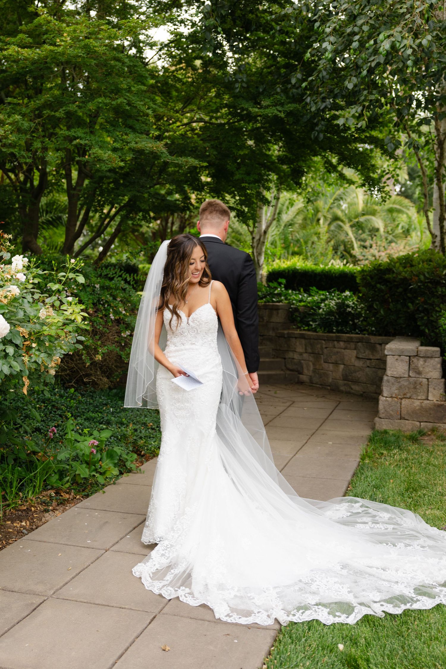 A wedding dress with an elegant lace train flows across a stone pathway in a lush garden setting.