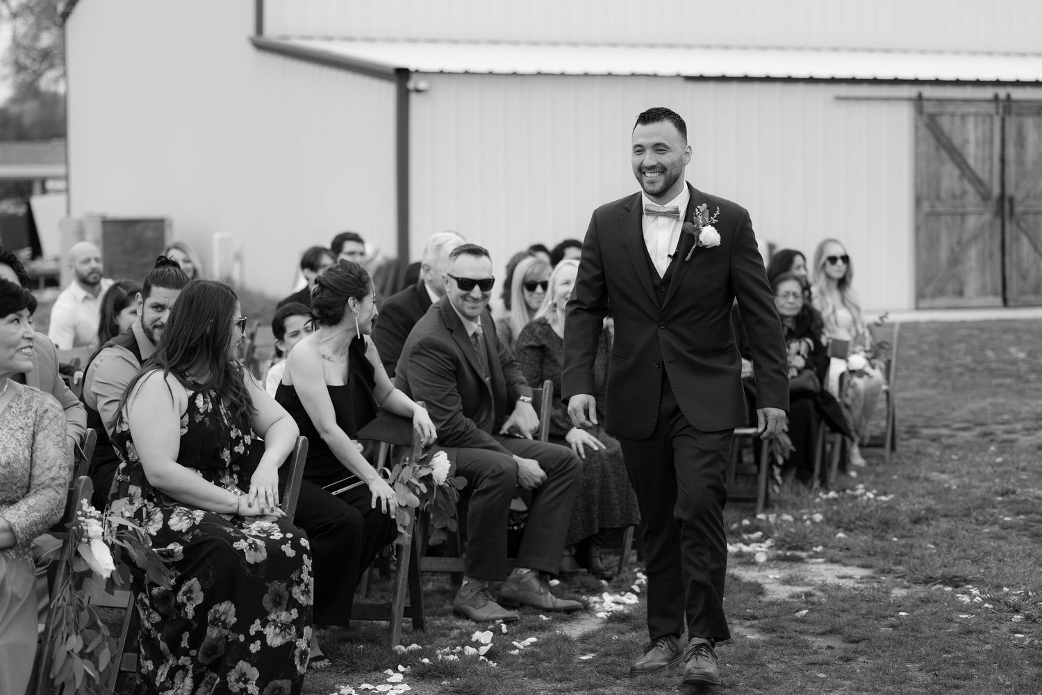 Black and white photo of an outdoor wedding ceremony with guests seated on chairs and someone standing in a suit.