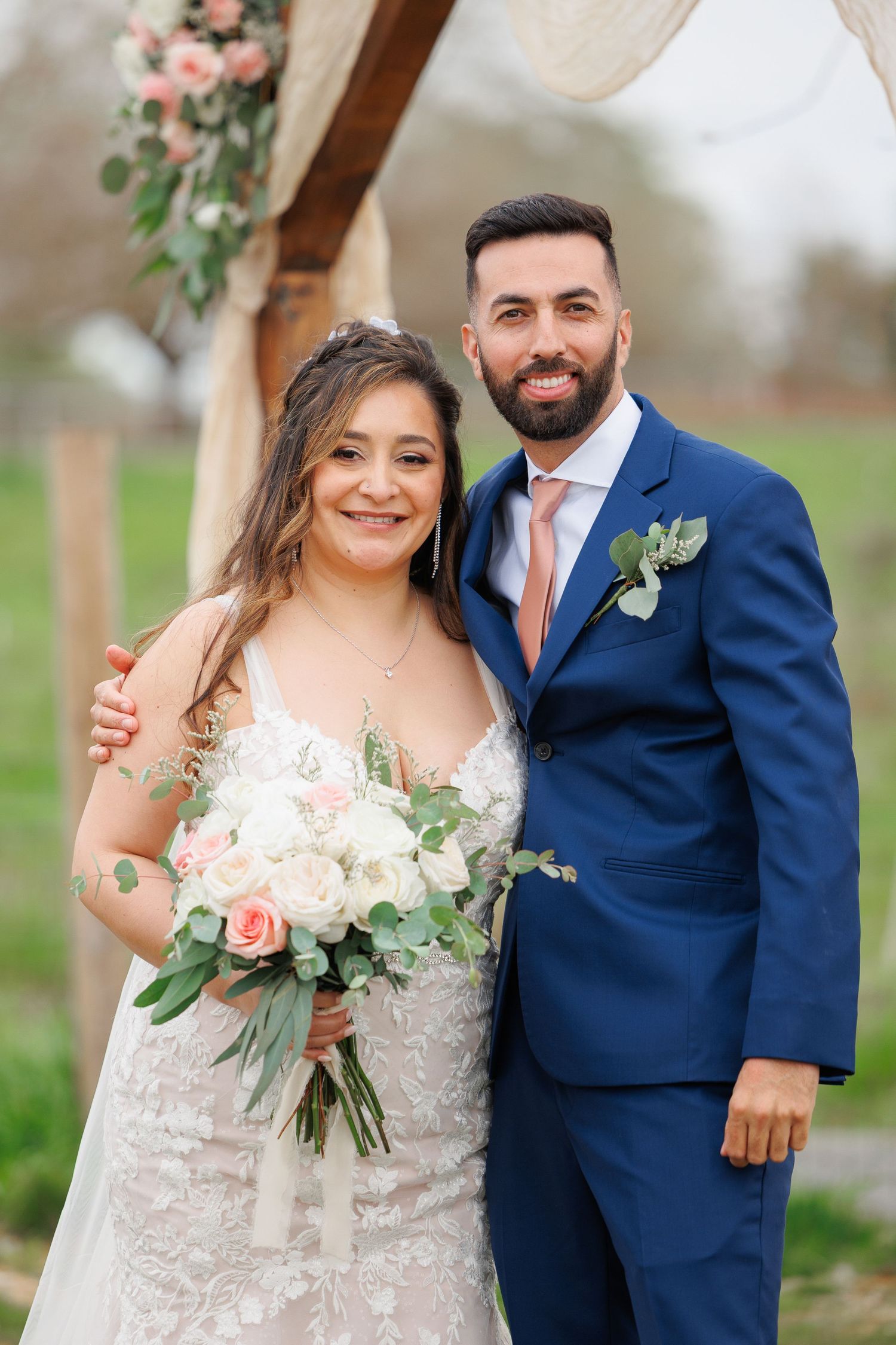 A couple poses together outdoors on their wedding day wearing a white dress and navy suit with pink floral bouquet details.