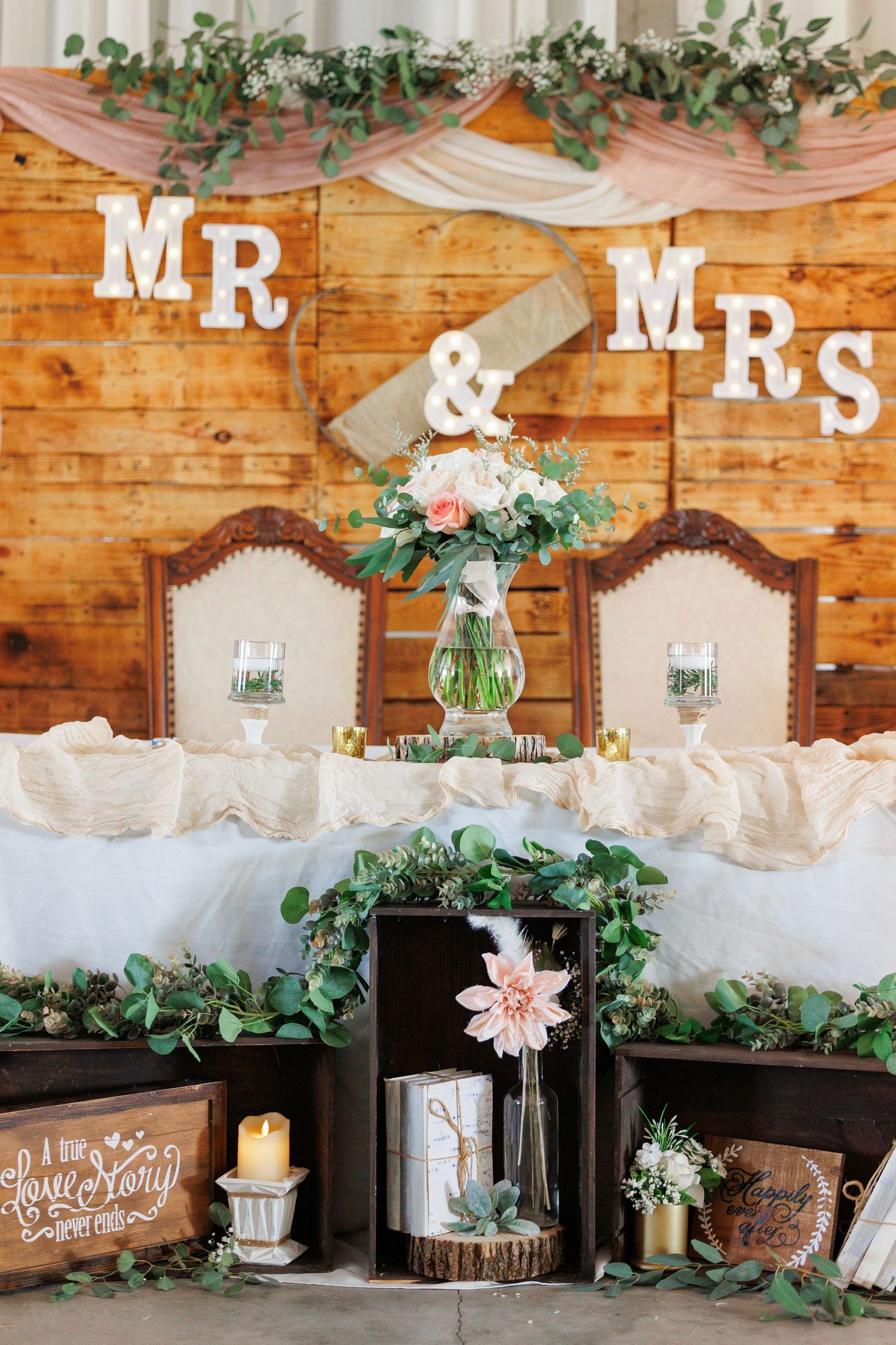 Rustic wedding decor with Mr & Mrs sign, draped fabric, greenery, and vintage crates on wooden backdrop.