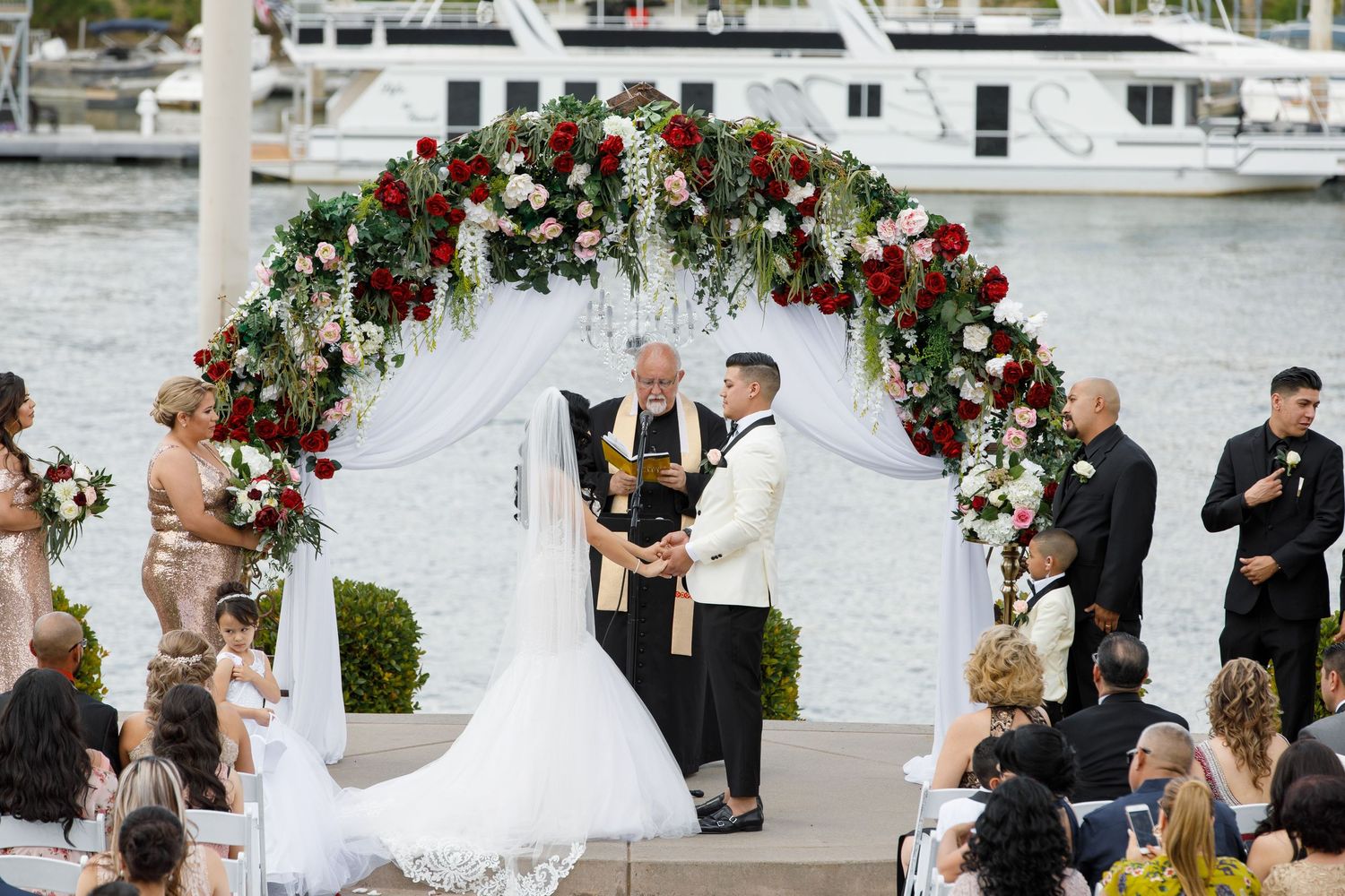 A wedding ceremony takes place outdoors at a marina with boats and floral decorations visible in the background.
