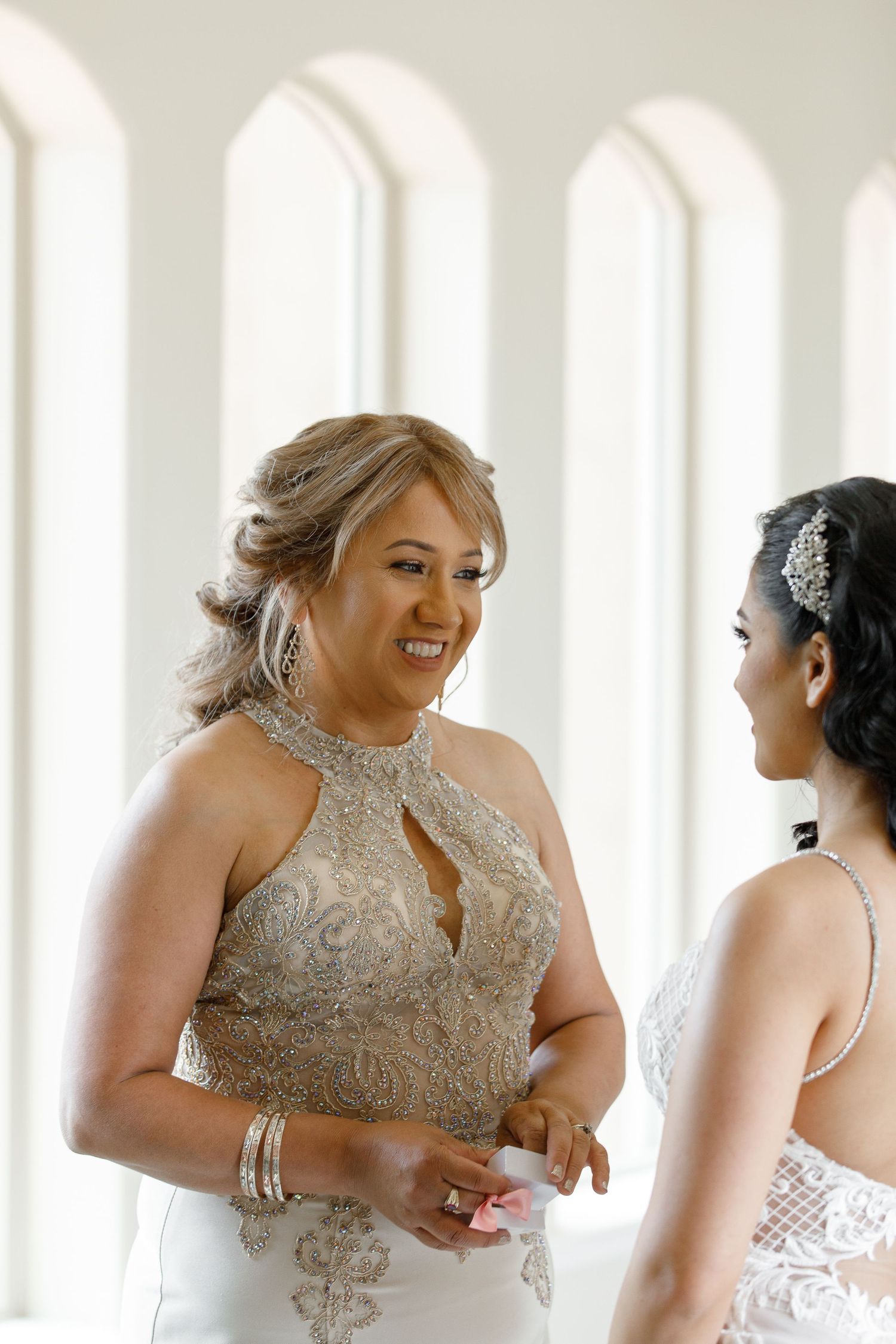 A bride and bridesmaid share a joyful moment while getting ready, both wearing elegant formal dresses in white and beige tones.