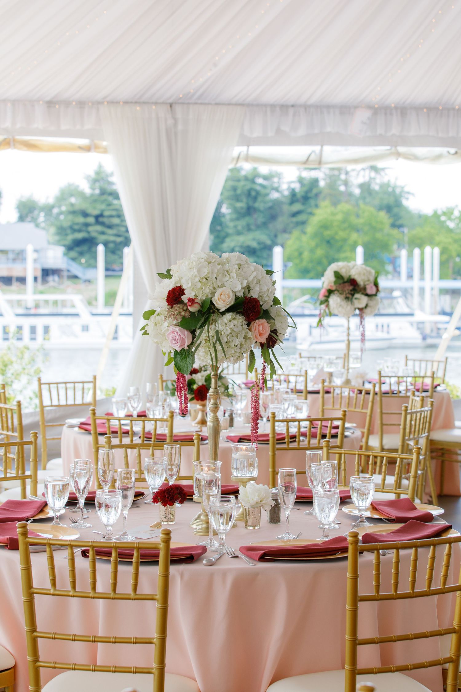 Elegant wedding reception tables set with burgundy accents, gold chiavari chairs and white floral centerpieces under white tent.