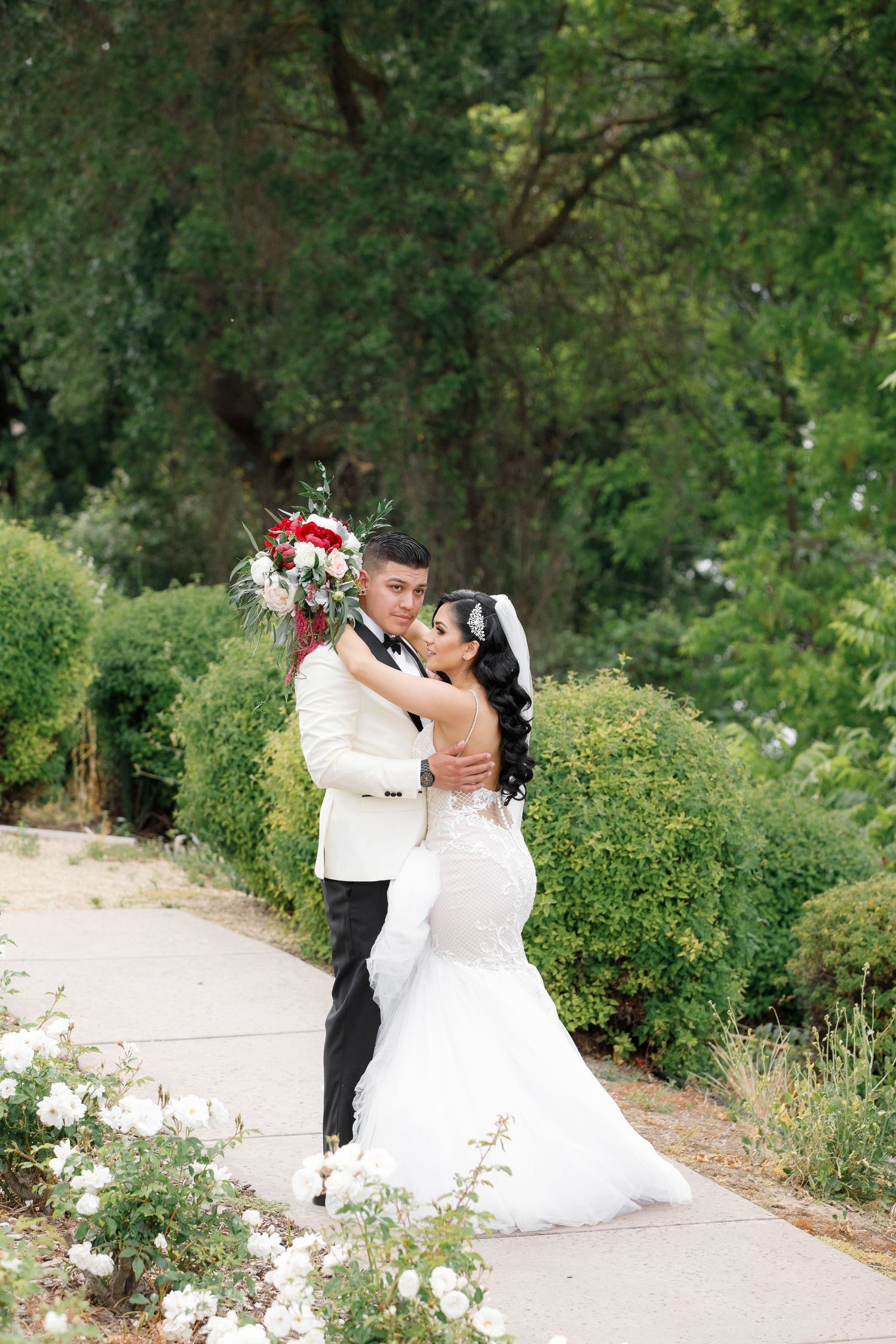 A romantic wedding couple dances together on a garden path surrounded by lush greenery and flowers.