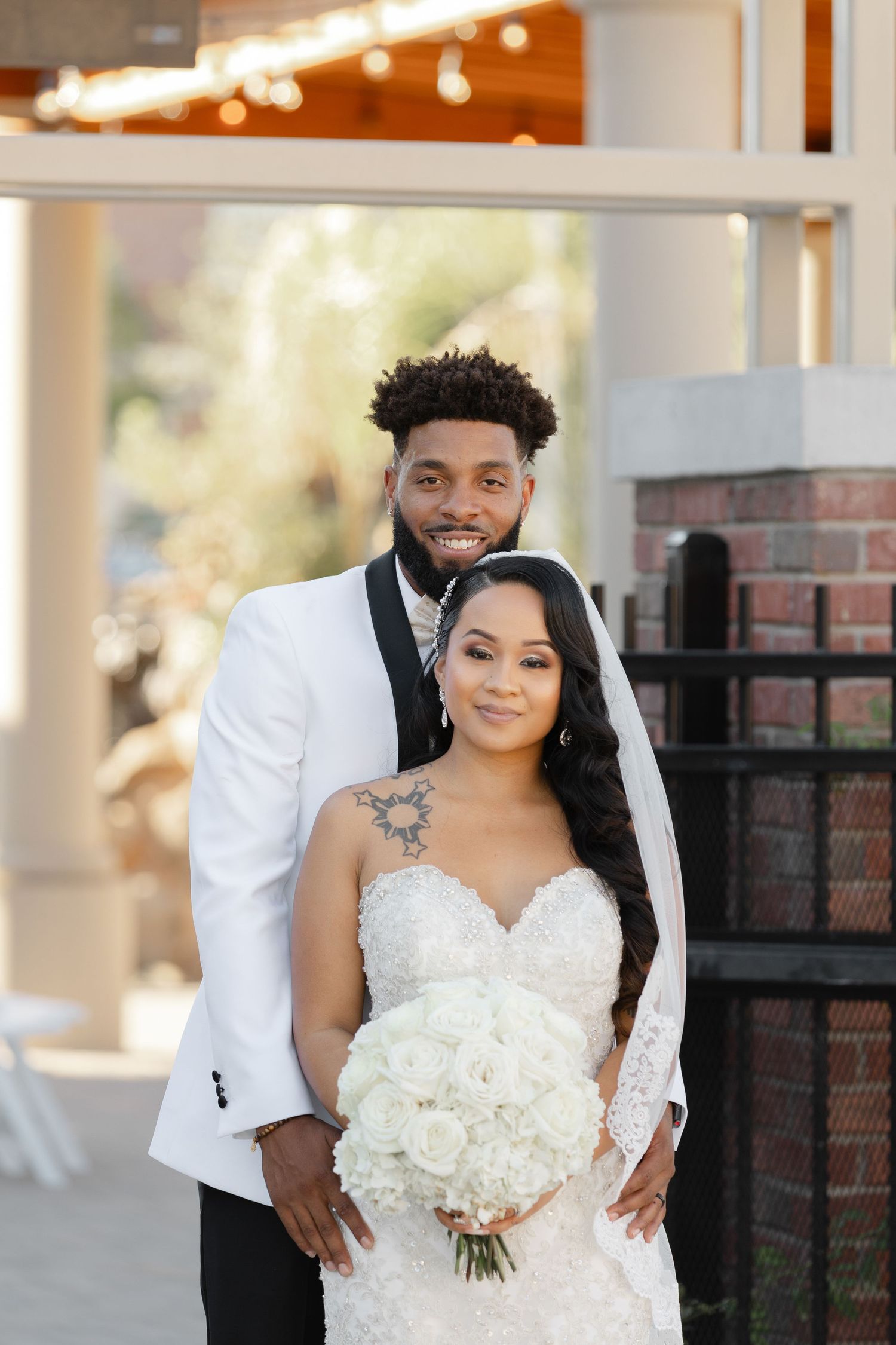 A couple poses together on their wedding day, with the bride holding a white bouquet and wearing a lace gown.