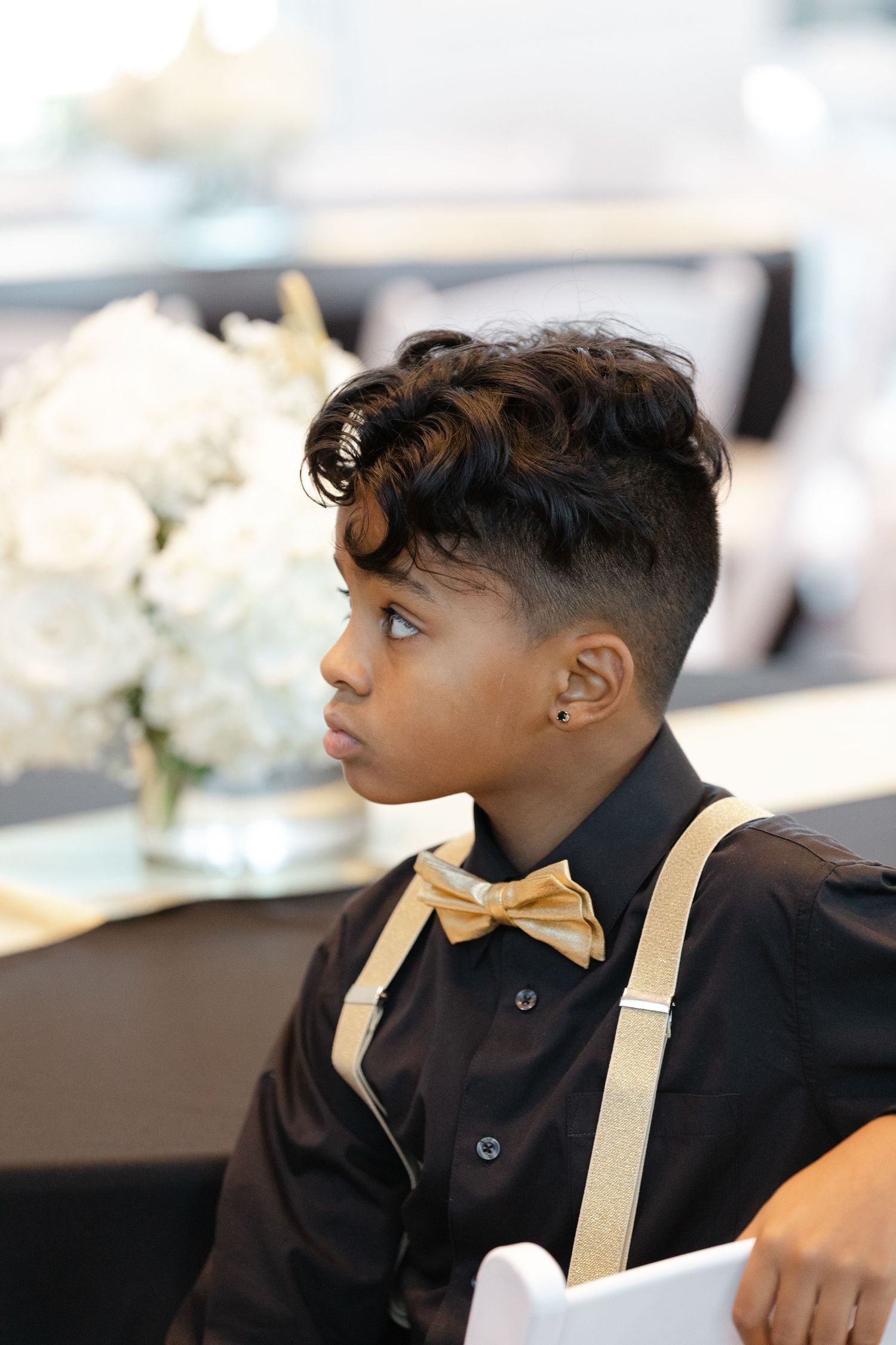 Young person in a black dress shirt with gold suspenders looking off to the side at a formal event.