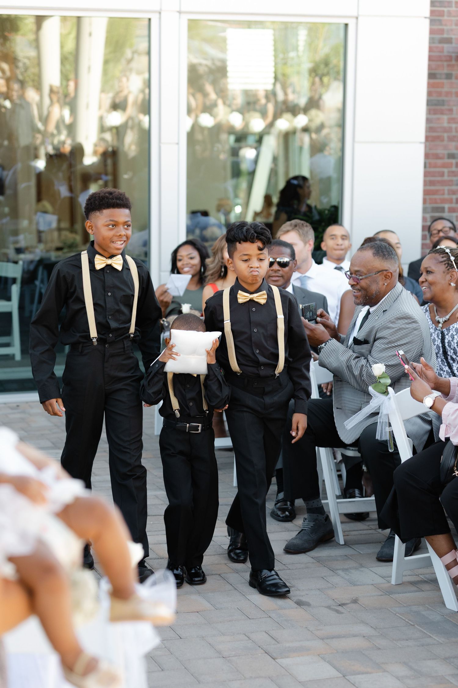 Young ring bearers in black suits with suspenders and bow ties walk down the aisle at an outdoor wedding ceremony.