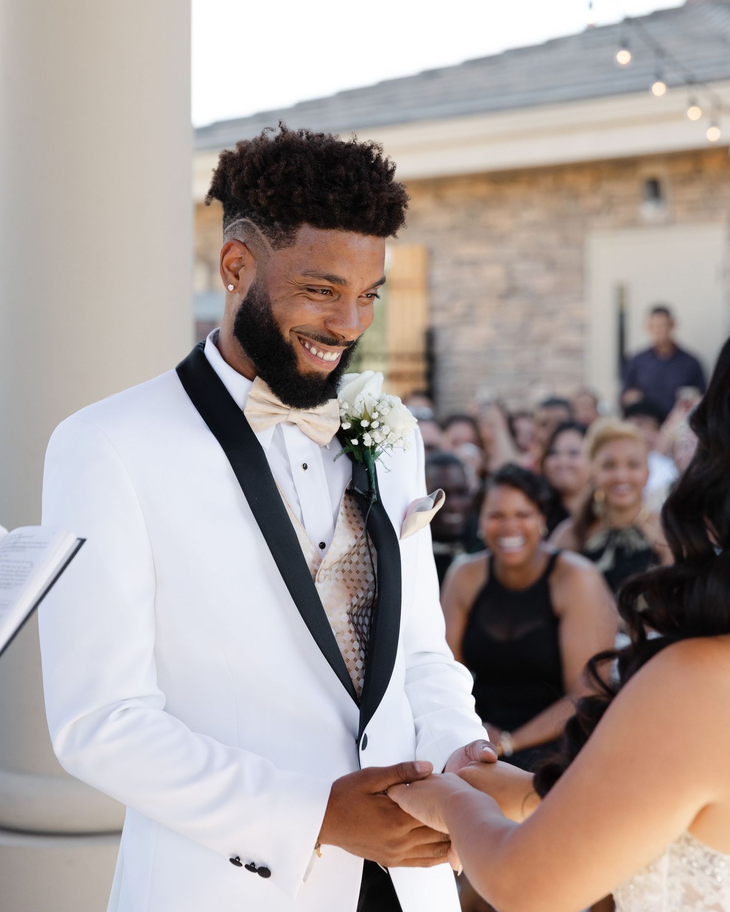 A groom in a white tuxedo jacket and black bowtie shares a joyful moment during an outdoor wedding ceremony.