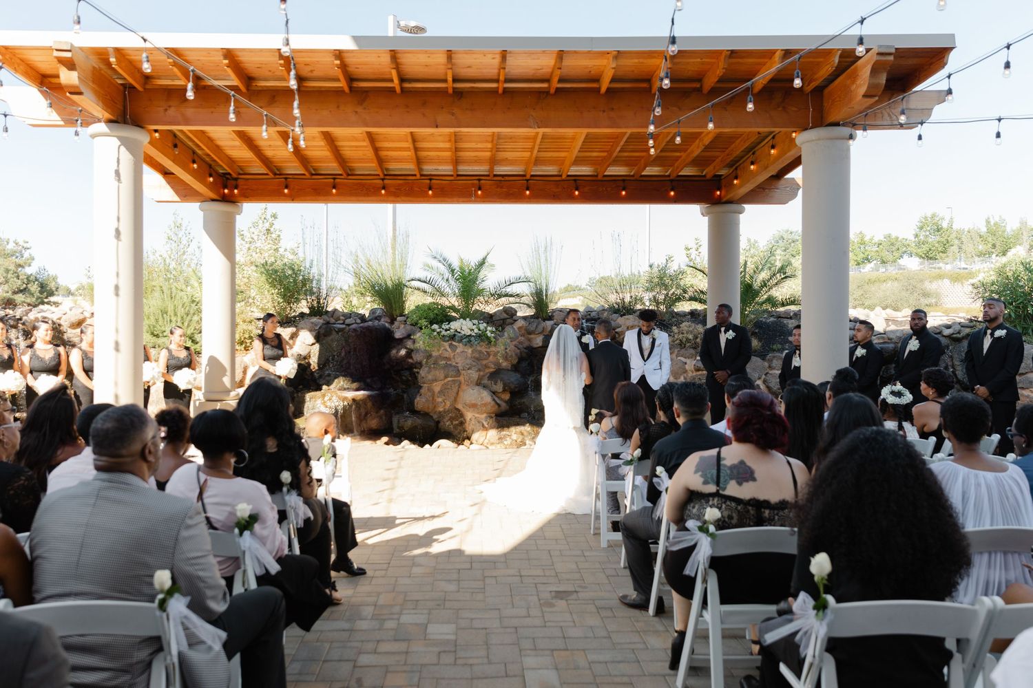 Panoramic view of an outdoor wedding ceremony with wooden pergola and guests seated in white chairs under a sunny sky.