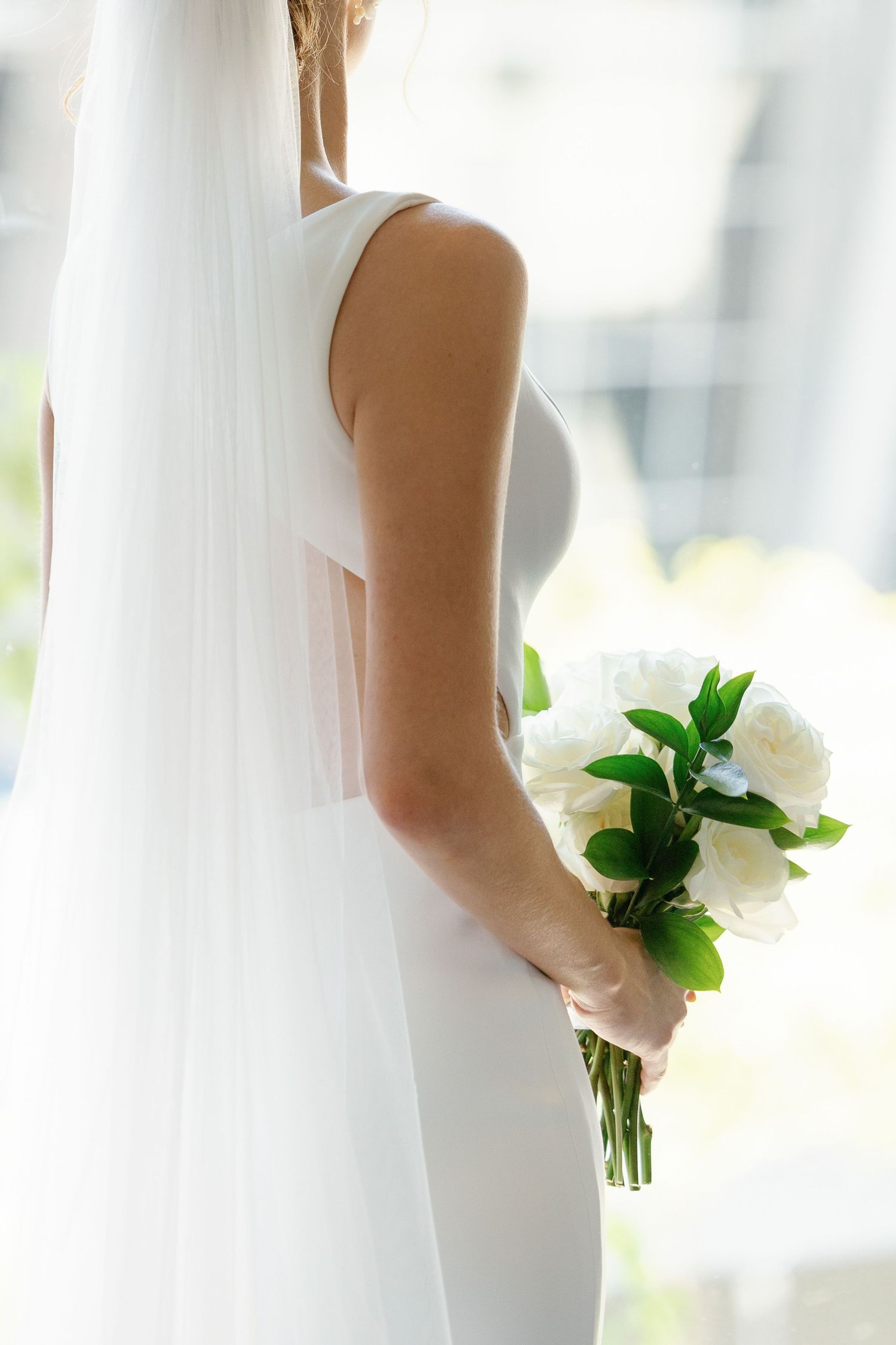A bride in a white wedding dress holds an elegant white floral bouquet against a bright, airy background.