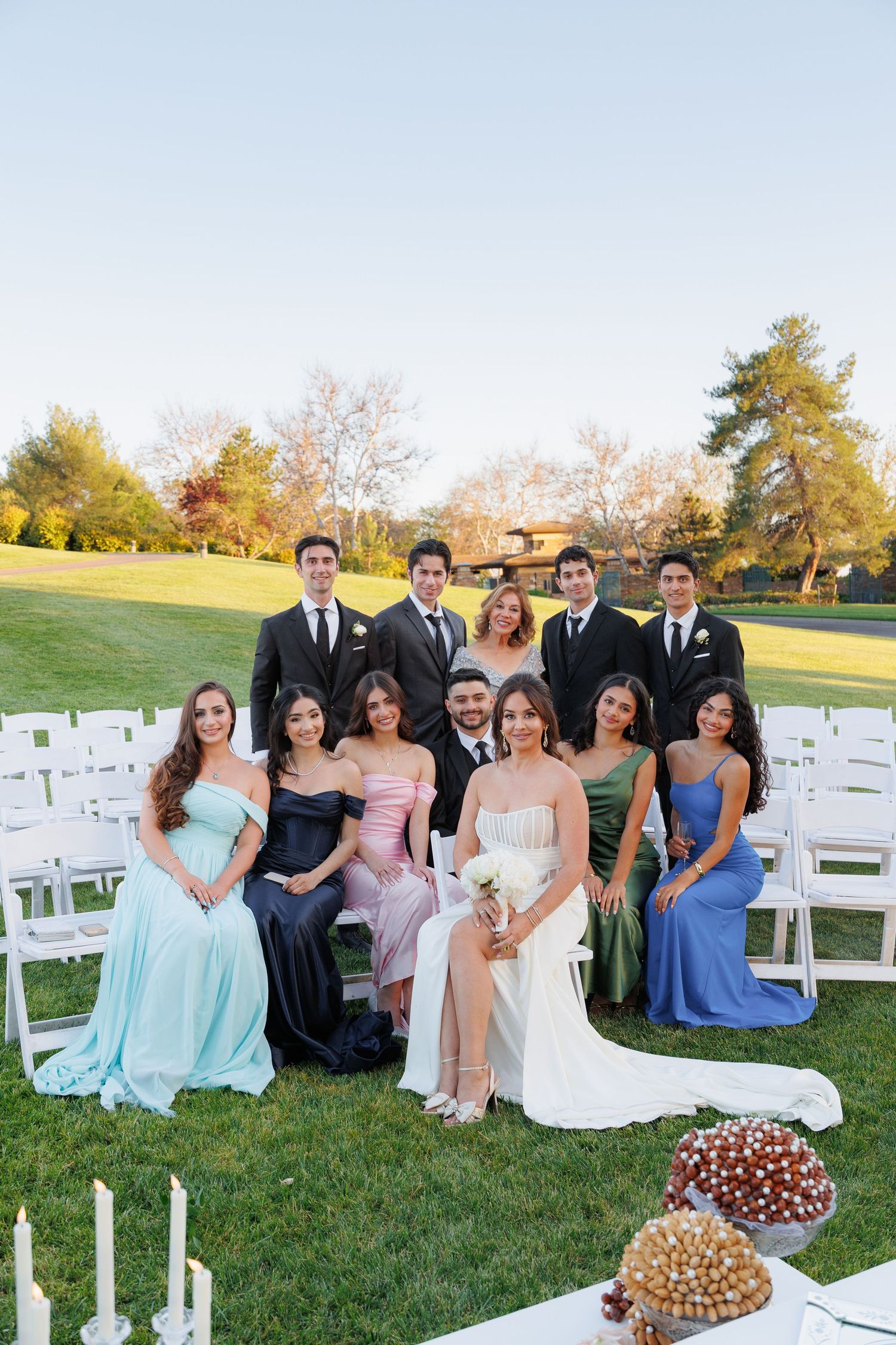 Wedding party poses on green lawn with white chairs and dessert displays at outdoor ceremony venue during sunset.