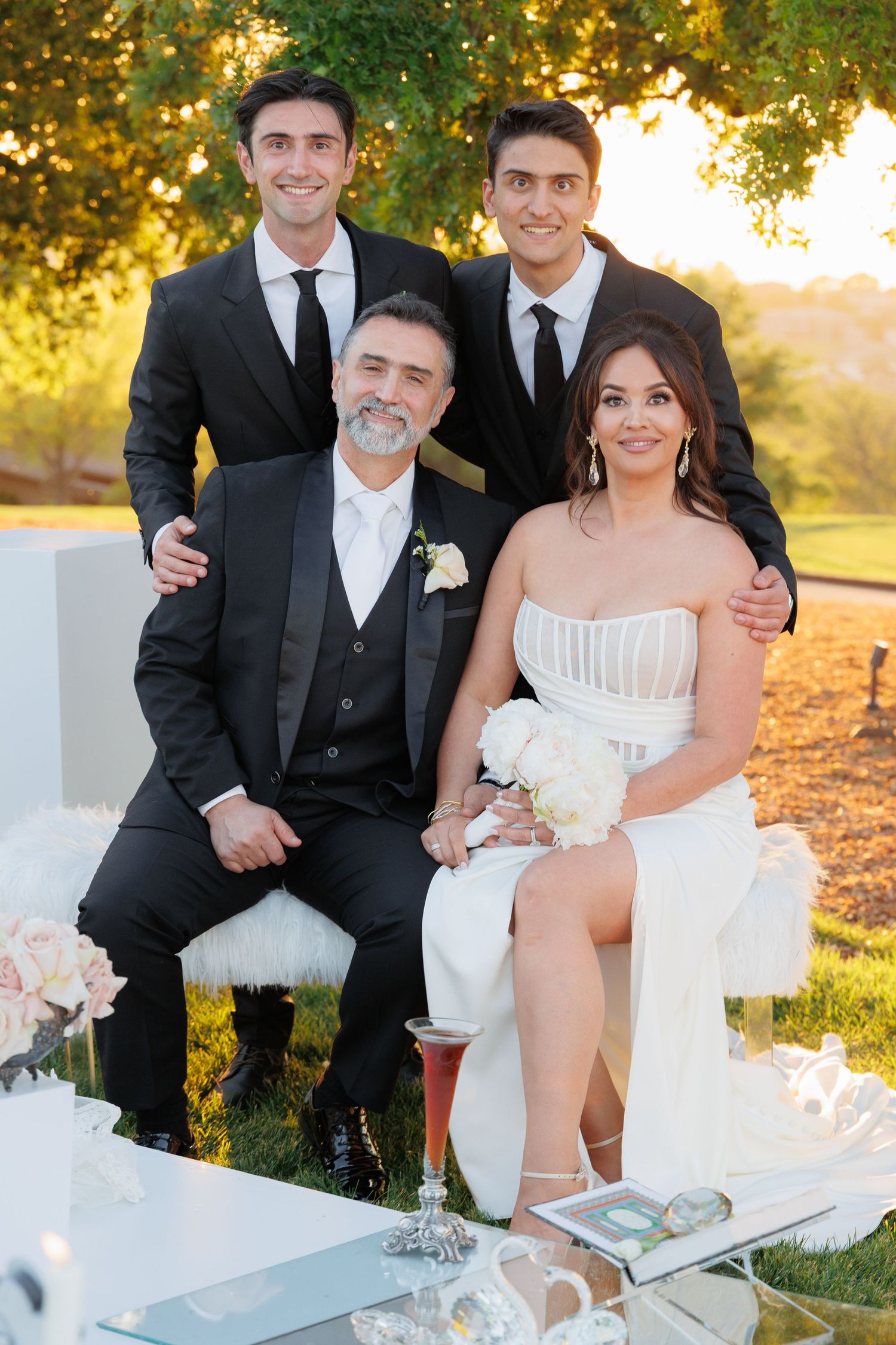 A formal family portrait outdoors at sunset with two young men standing behind a seated couple in wedding attire.