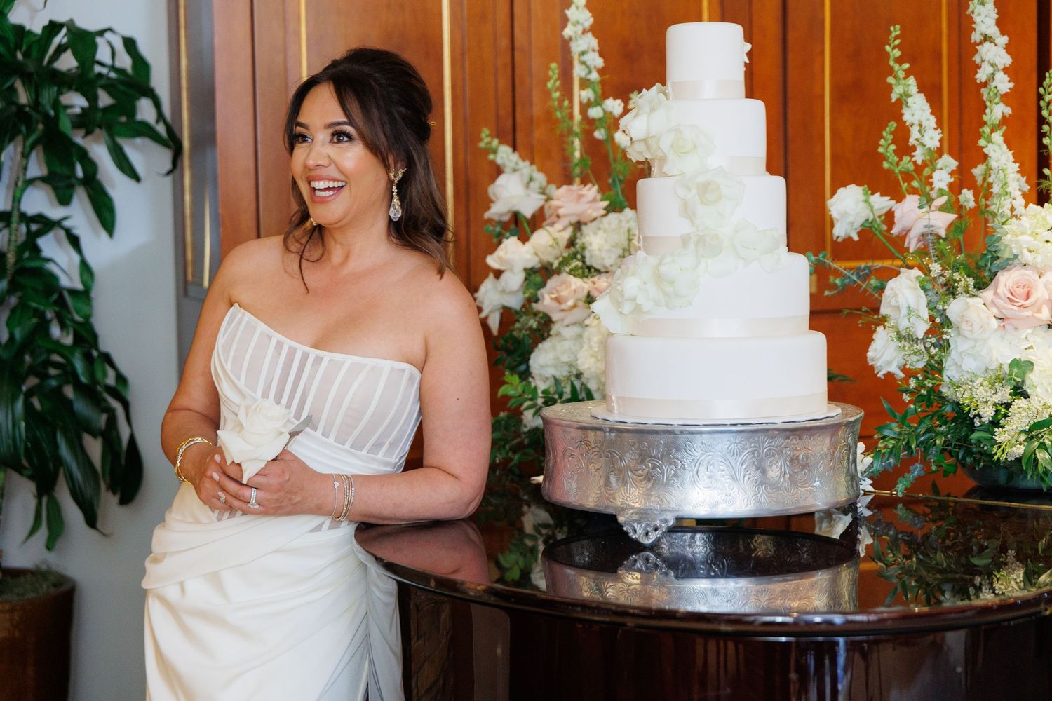 Elegant white wedding cake with delicate floral details sits on a dark wooden table at a reception venue.