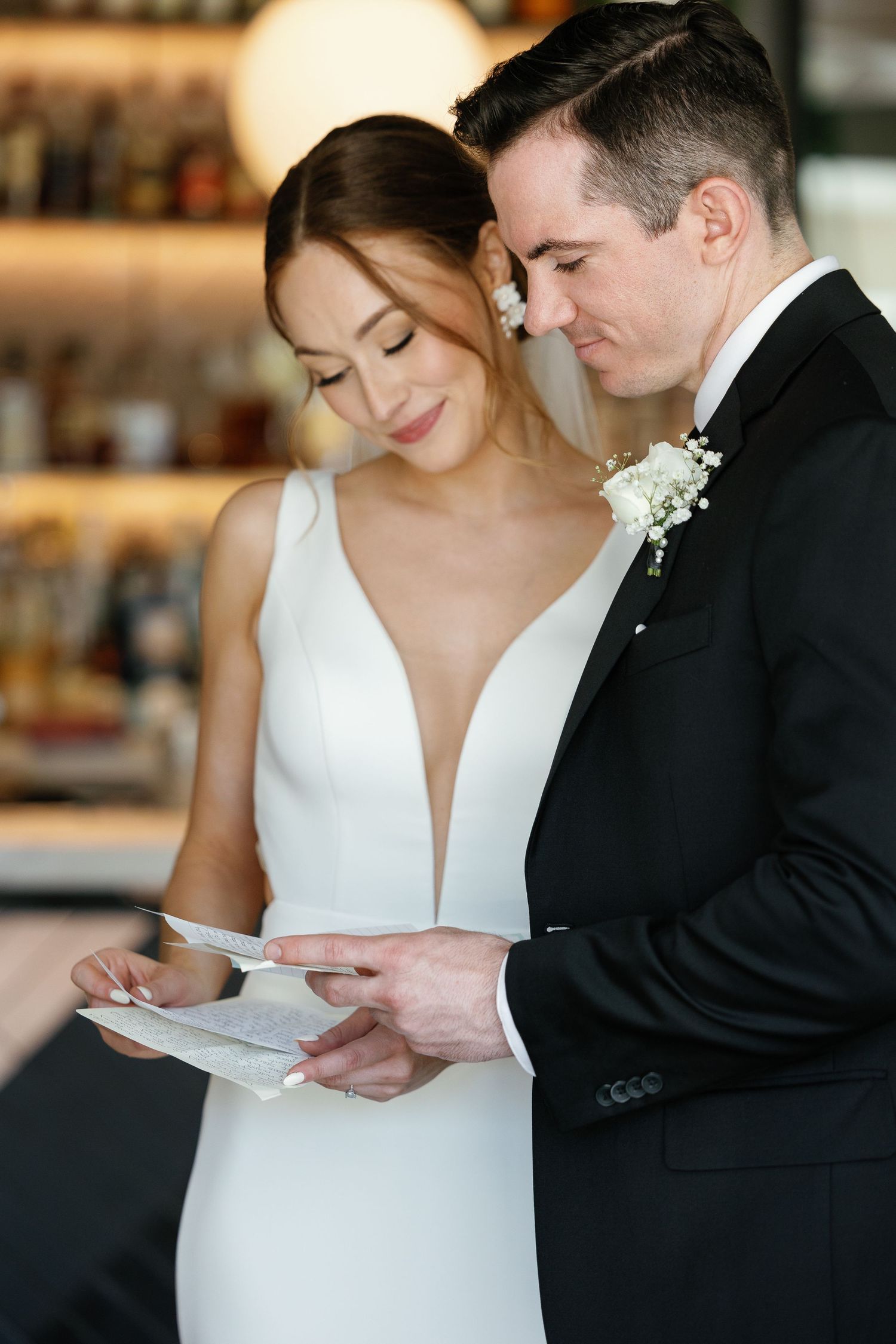 A bride in white gown and groom in black suit read a letter together at their wedding.